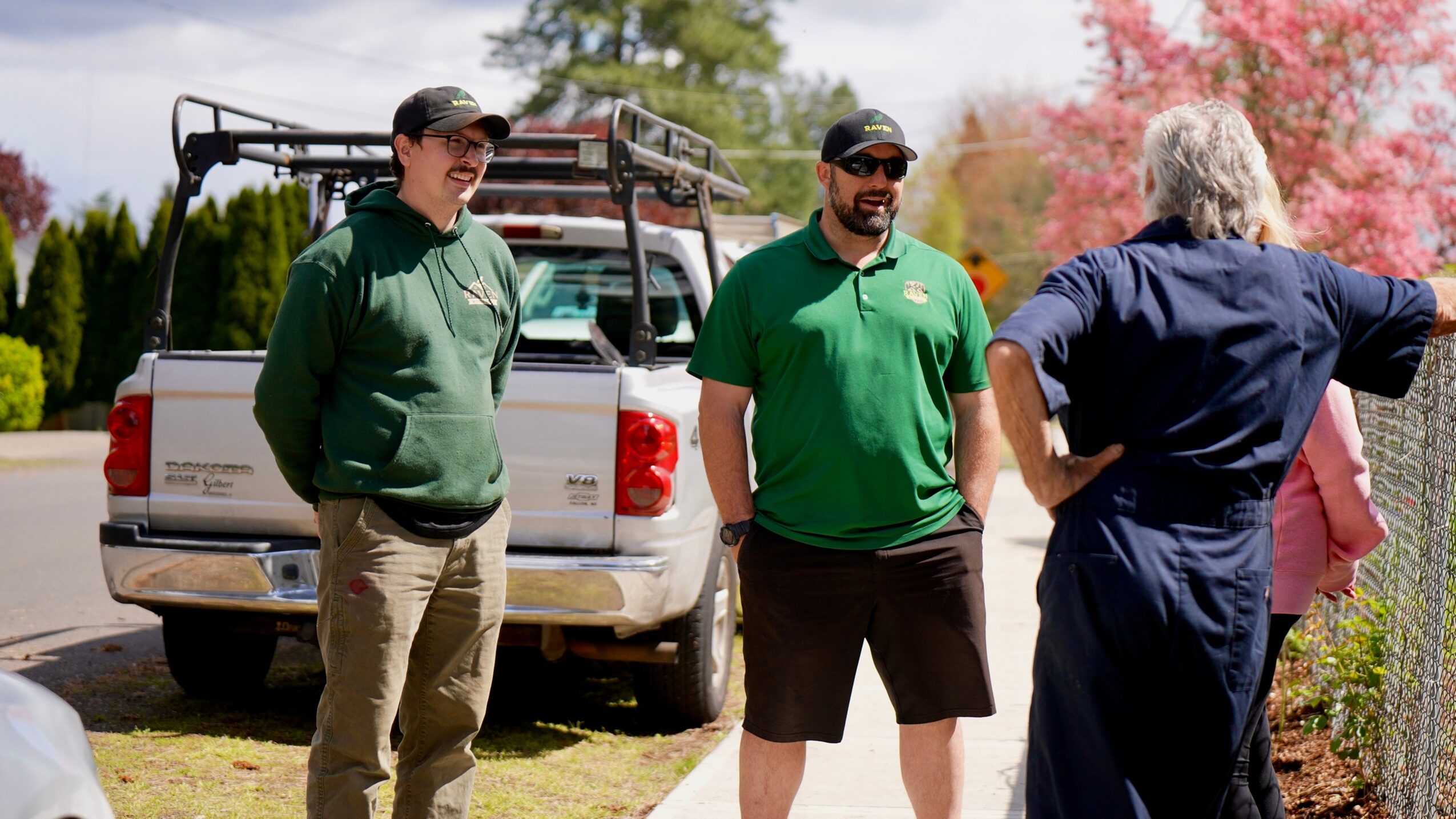 Three men standing outdoors near a white vehicle, engaged in conversation, with trees and pink blossoms in the background.