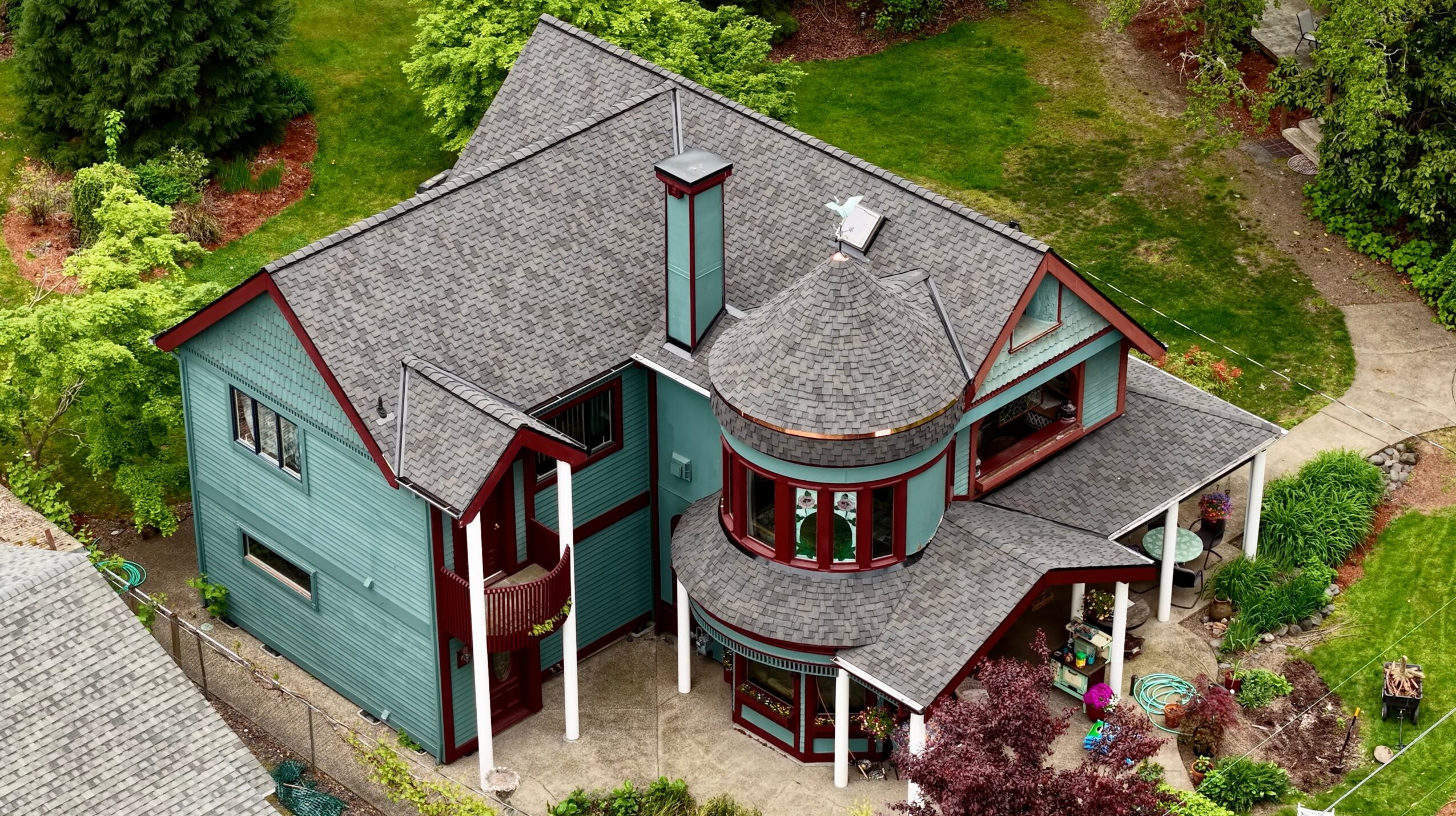 A large house with multiple roof sections, a chimney, and a wrap-around porch, surrounded by trees and greenery.