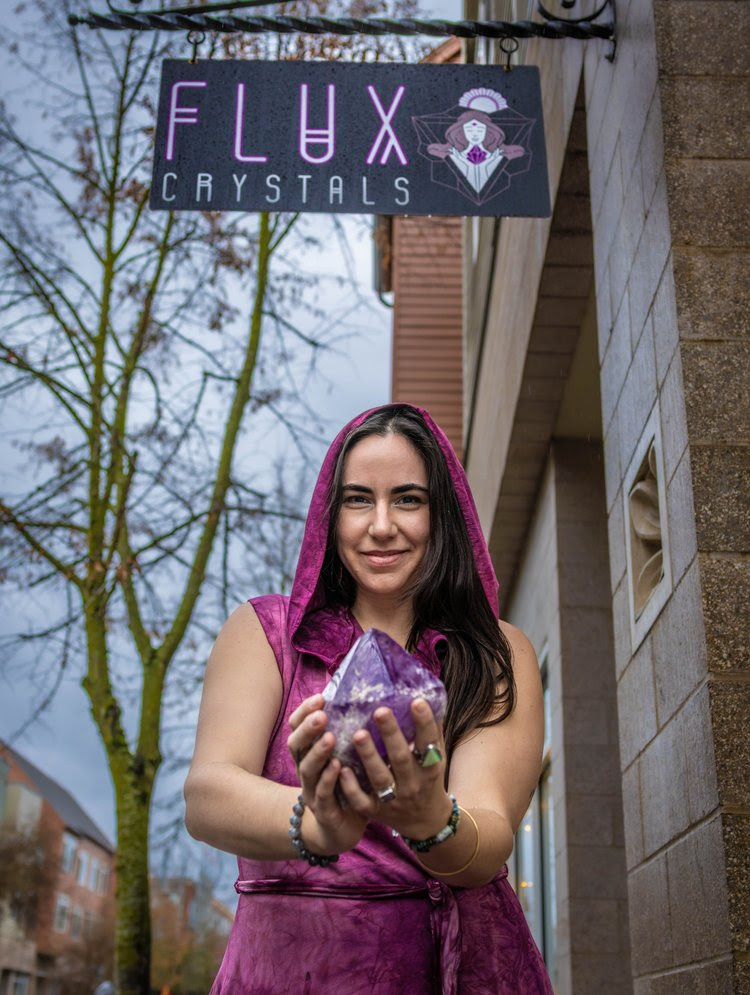 Woman holding a large purple crystal in front of a store sign that reads 'FLUX CRYSTALS'.