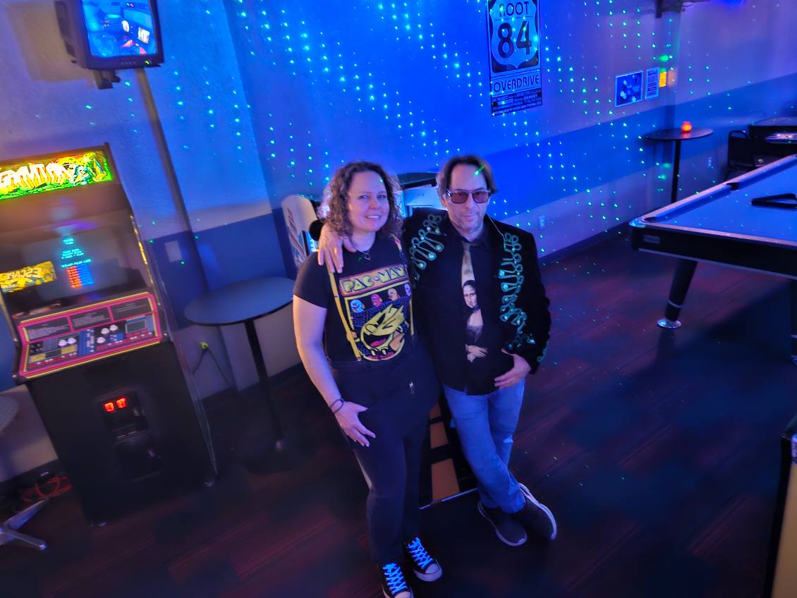 Two people standing in a room with blue lighting and a pool table, smiling at the camera.