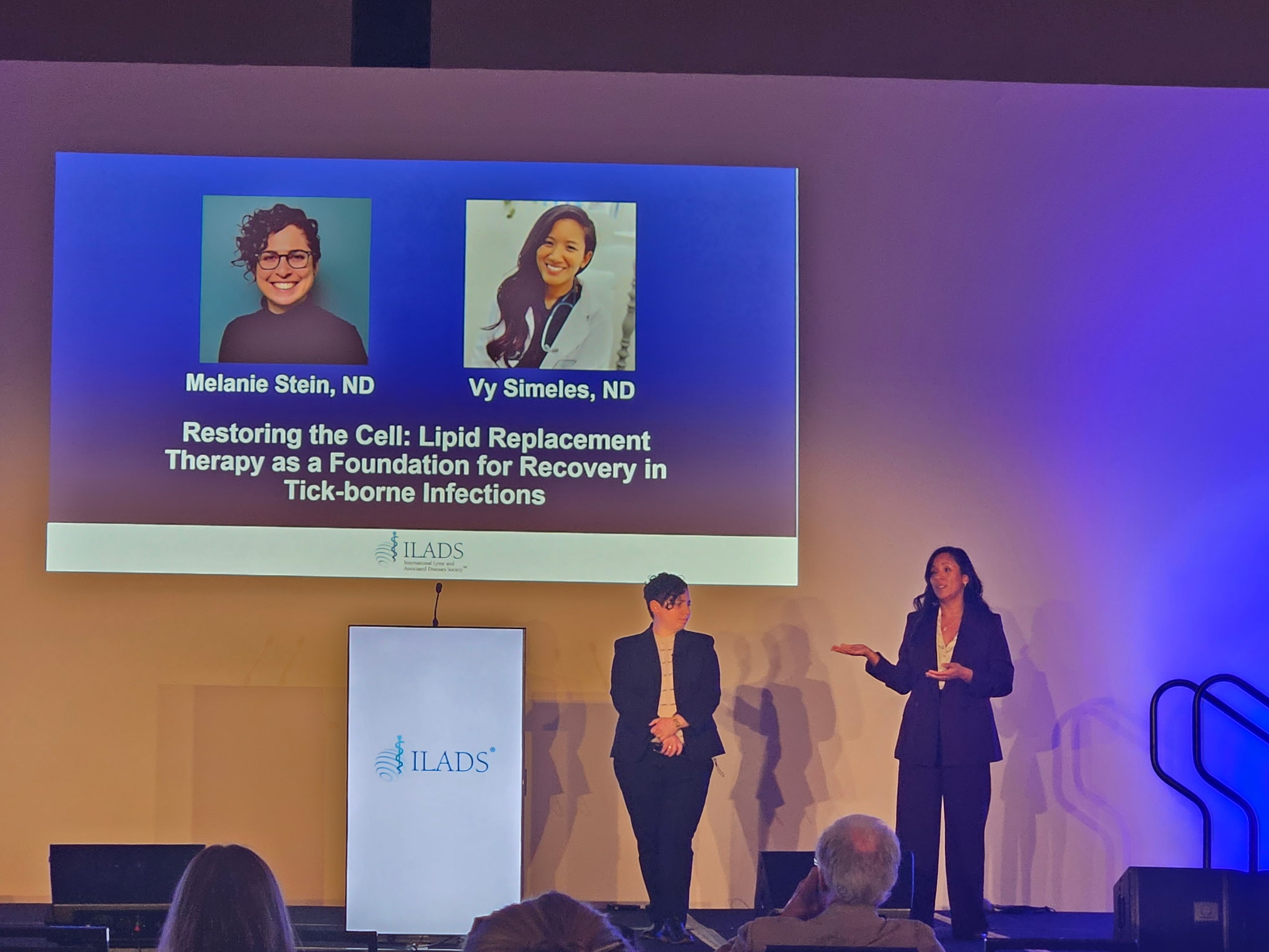 Two women standing on stage in front of a screen displaying presentation slides, with an audience in front.