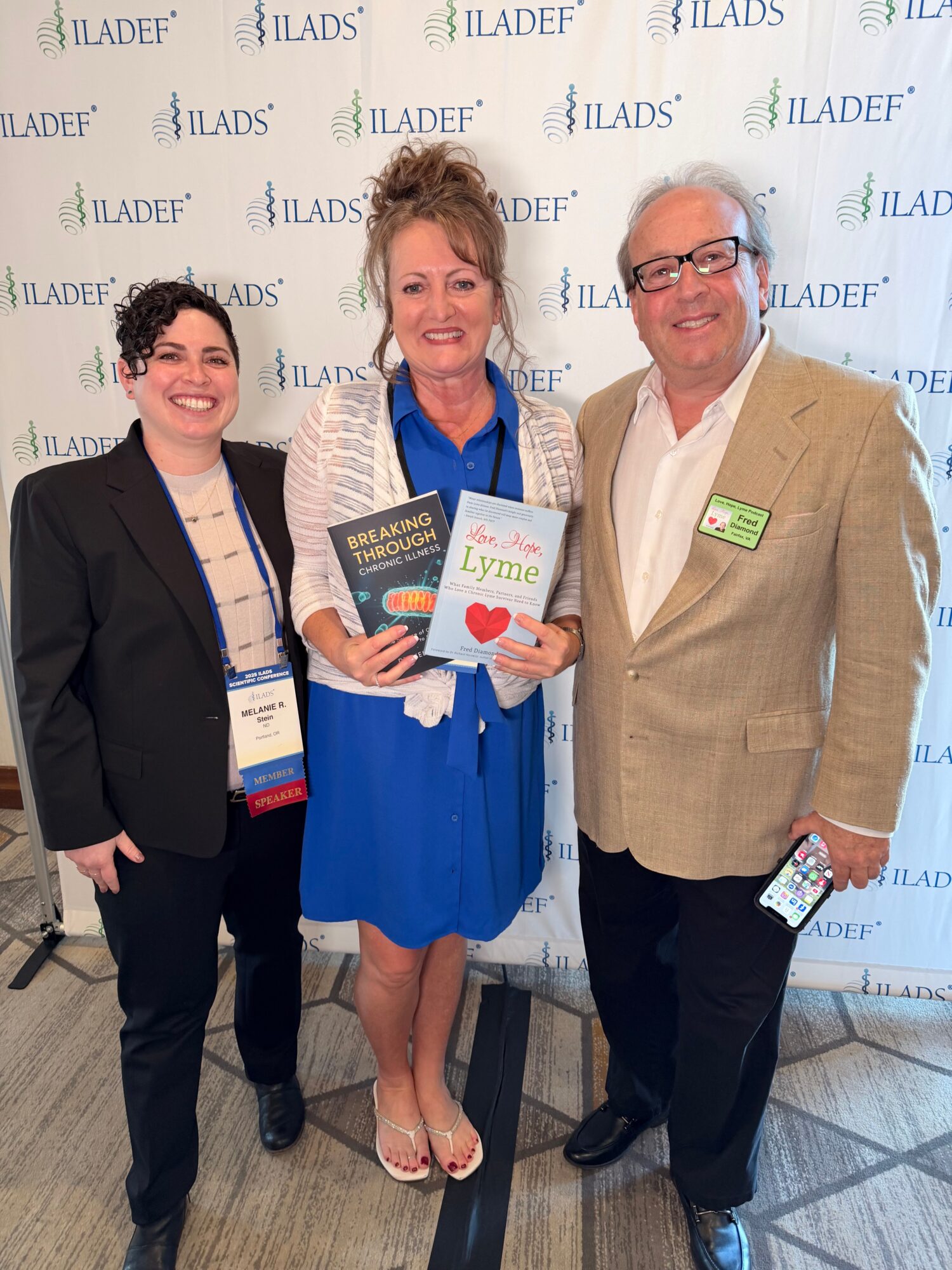 Three people standing together in front of a backdrop with ILADef logo, smiling, holding a book and a pamphlet.