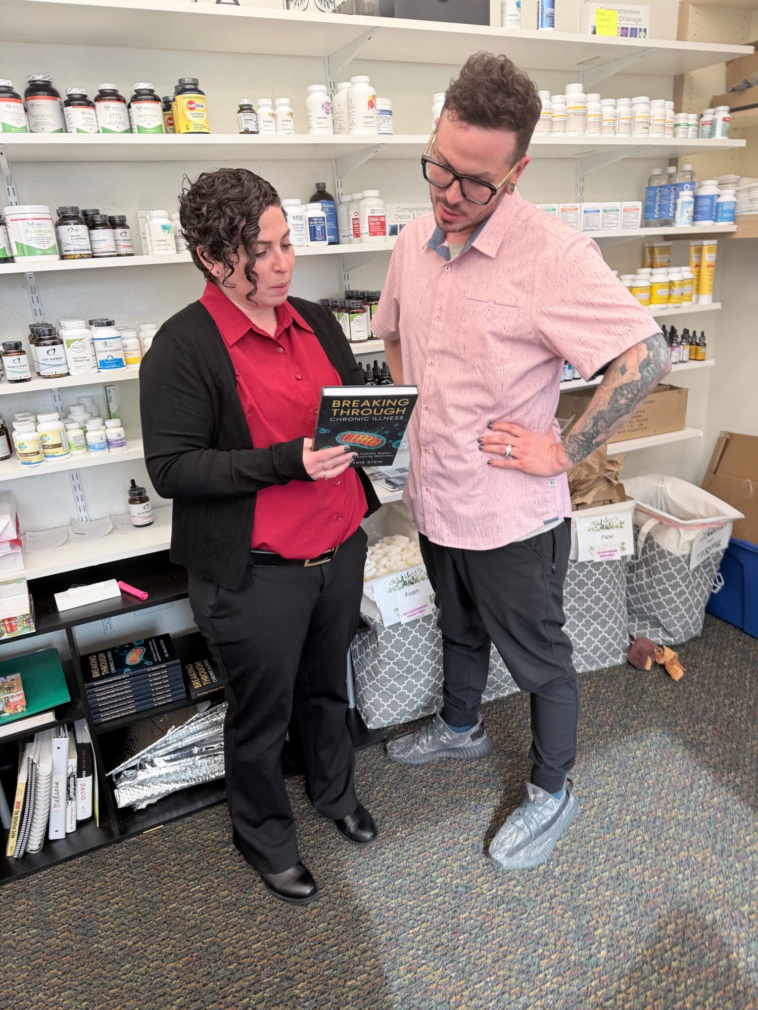 Two people standing in a pharmacy, one holding a tablet, shelves with medicines in the background.