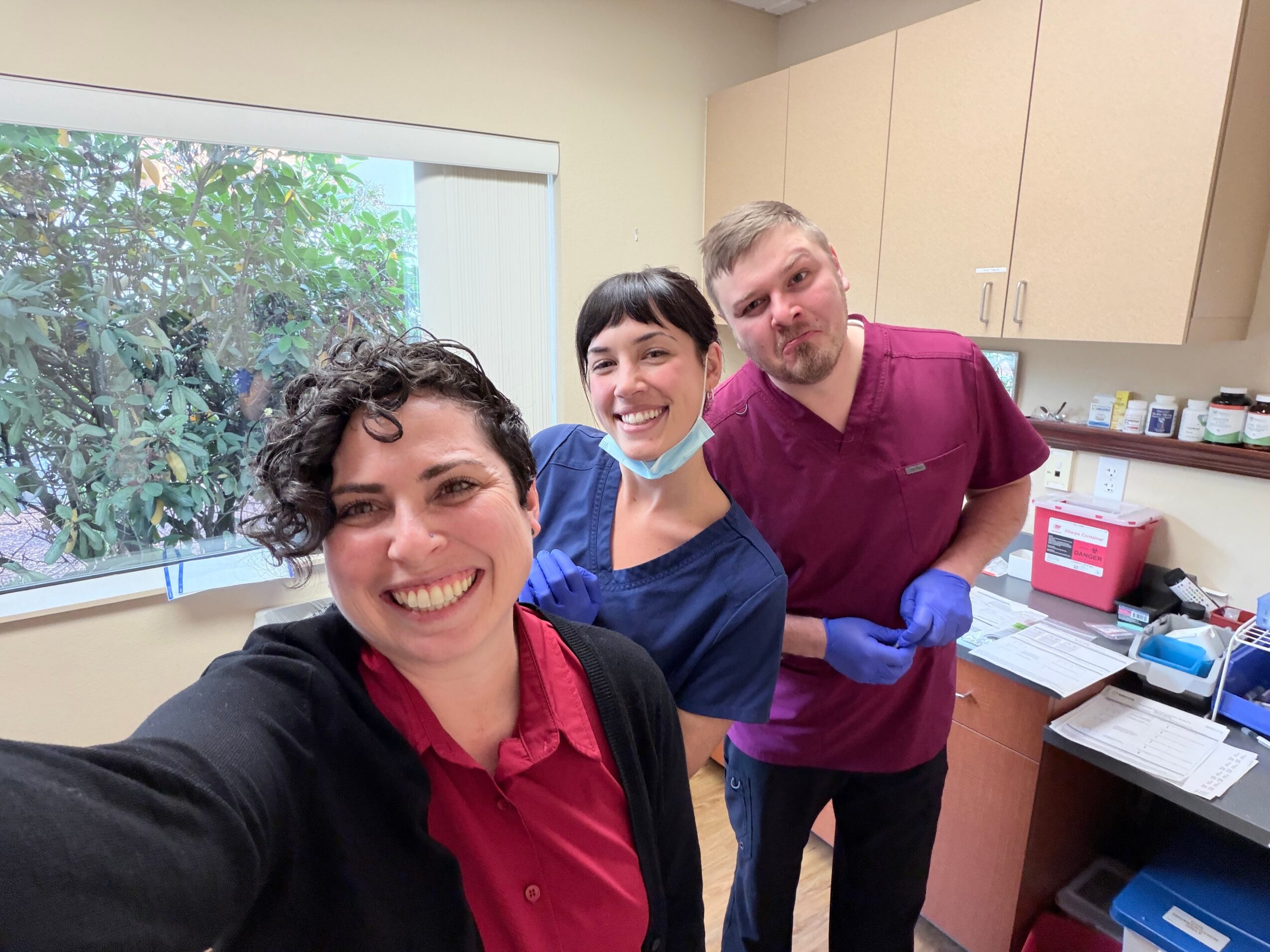 Three healthcare workers smiling in a clinic room, one woman in a red shirt, two others in scrubs, with a window and cabinets behind them.