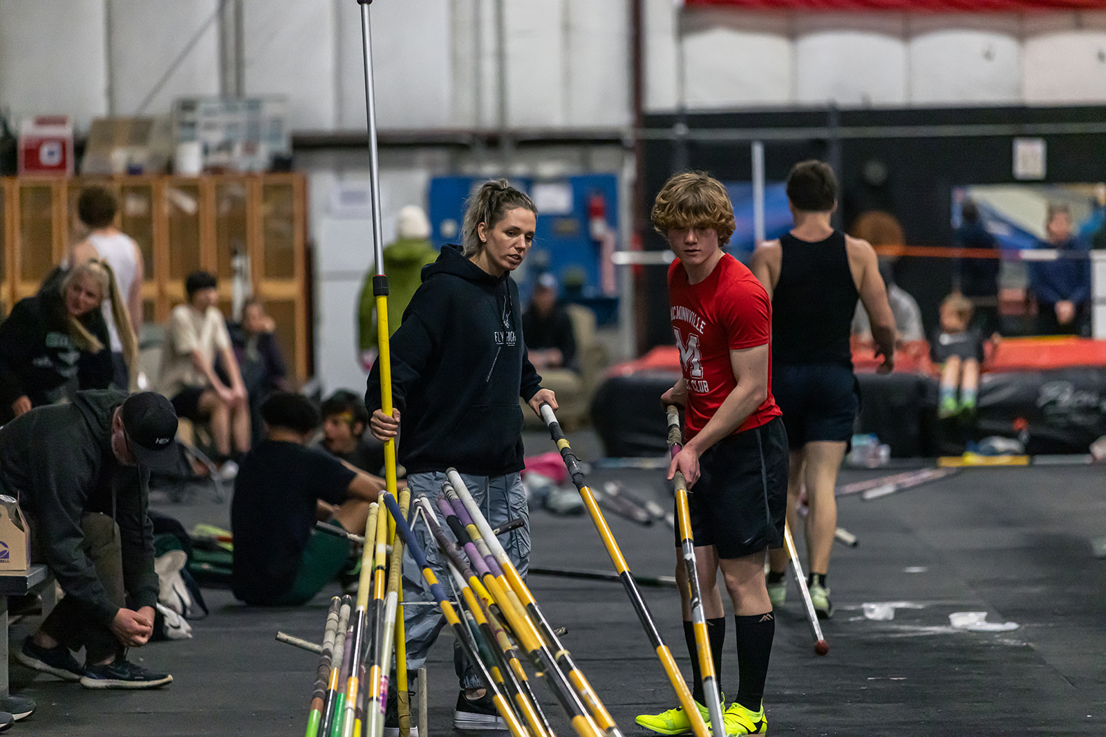 Indoor sports facility with people preparing for pole vaulting, some sitting and others standing, equipment visible in background.
