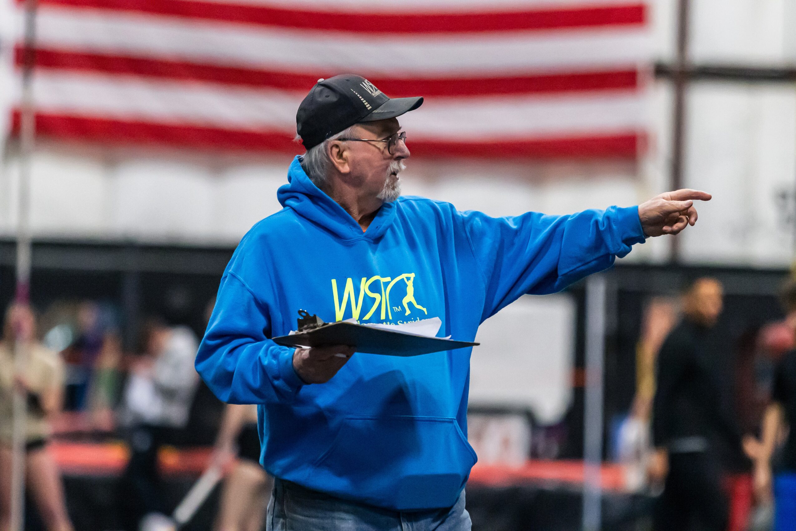 Older man in a blue hoodie and black cap pointing with his right arm, holding a clipboard, in an indoor setting with a blurred crowd and American flag in background.