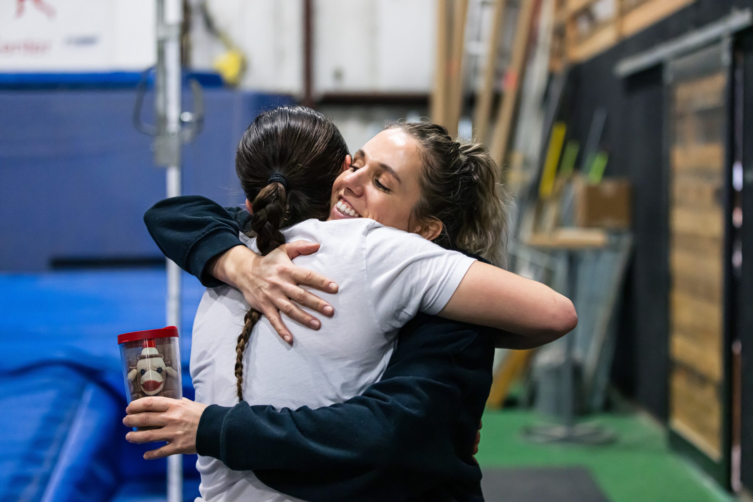 Two women hugging warmly in a workshop or industrial setting, one holding a cup with a bear design.