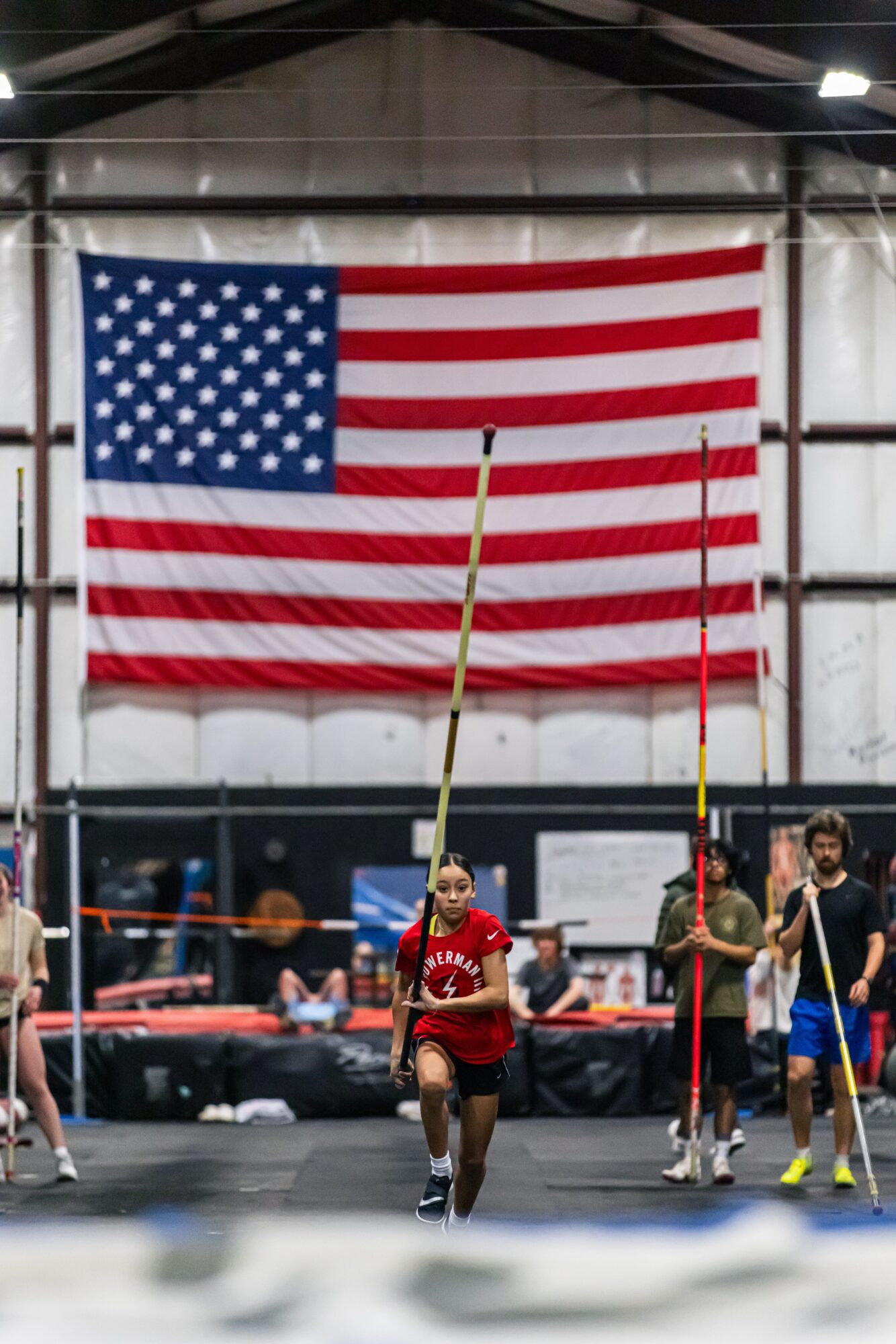 Indoor track and field event with athletes jumping over a pole, American flag hanging in the background.