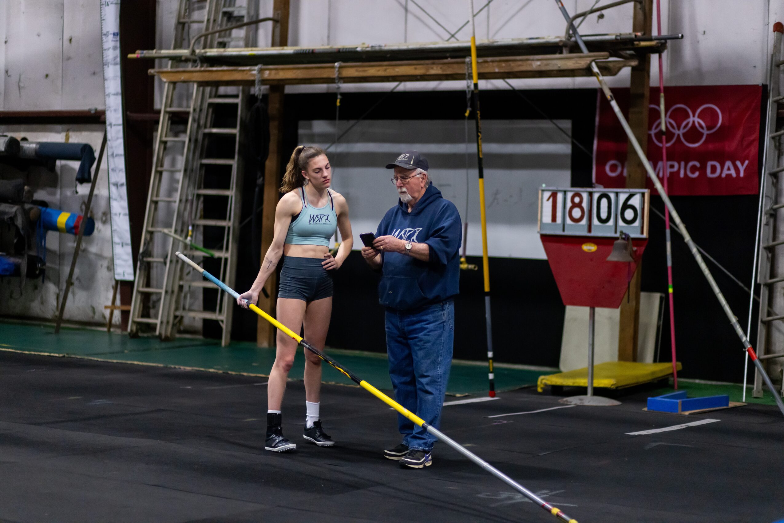 Two people standing on a track, one holding a clipboard, with a scoreboard showing 18.06 seconds, in an indoor sports facility.