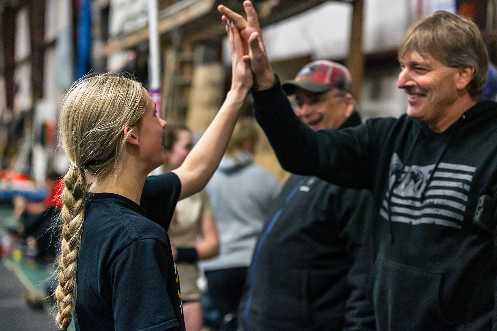A young girl and an older man high-five outdoors, smiling, with a crowd and market stalls in the background.