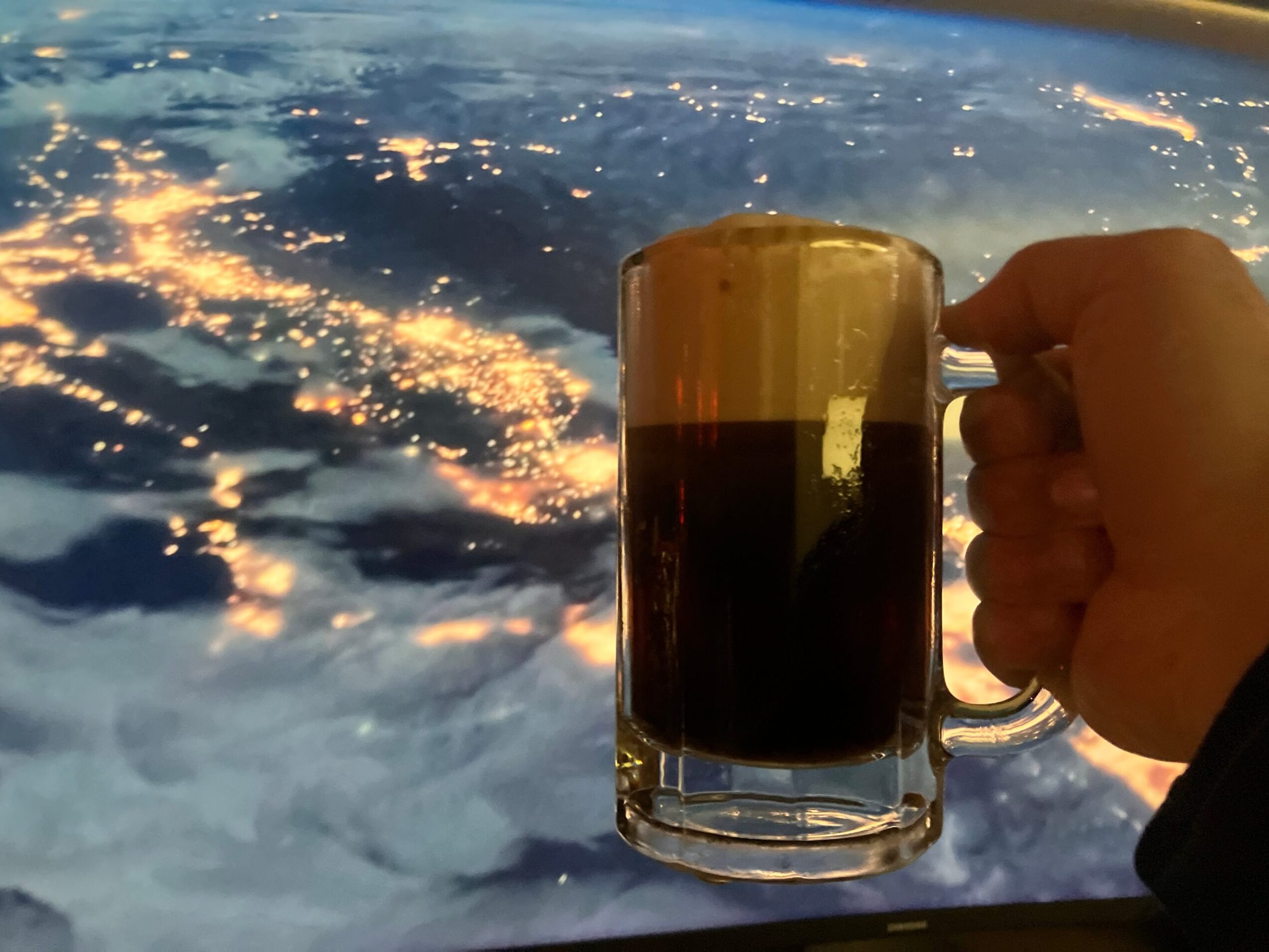 Person holding a glass mug of dark beverage against a cloudy sky with sunlight reflections.