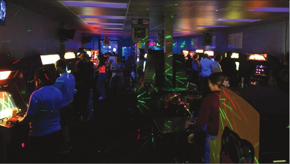 People playing arcade games in a dimly lit room with colorful lights and screens, some standing and some sitting.