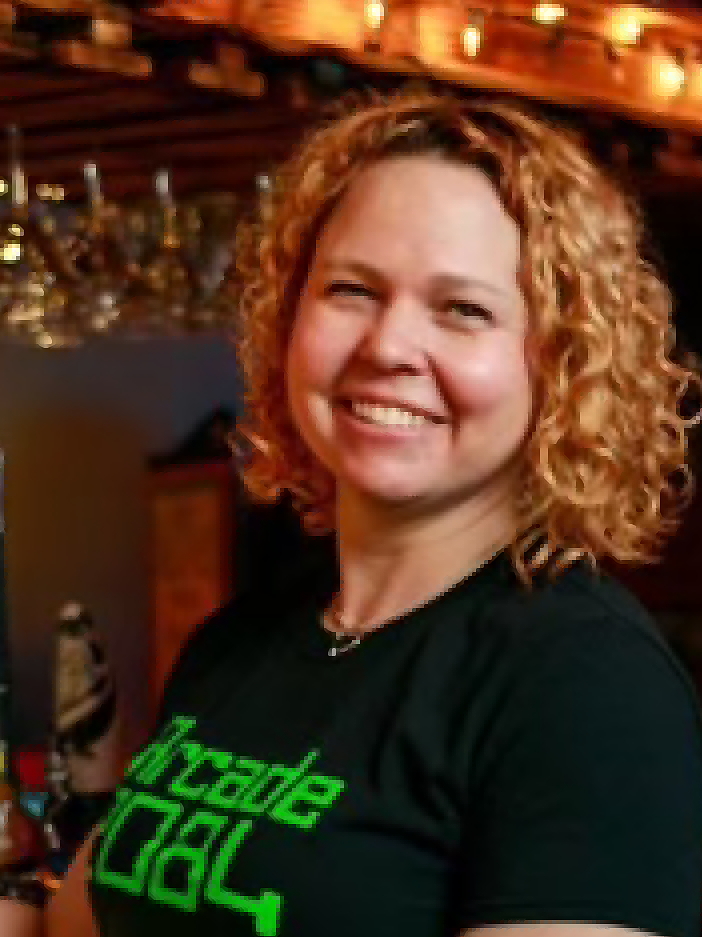 Smiling woman with curly hair in a black shirt with green text, in a warmly lit indoor setting.