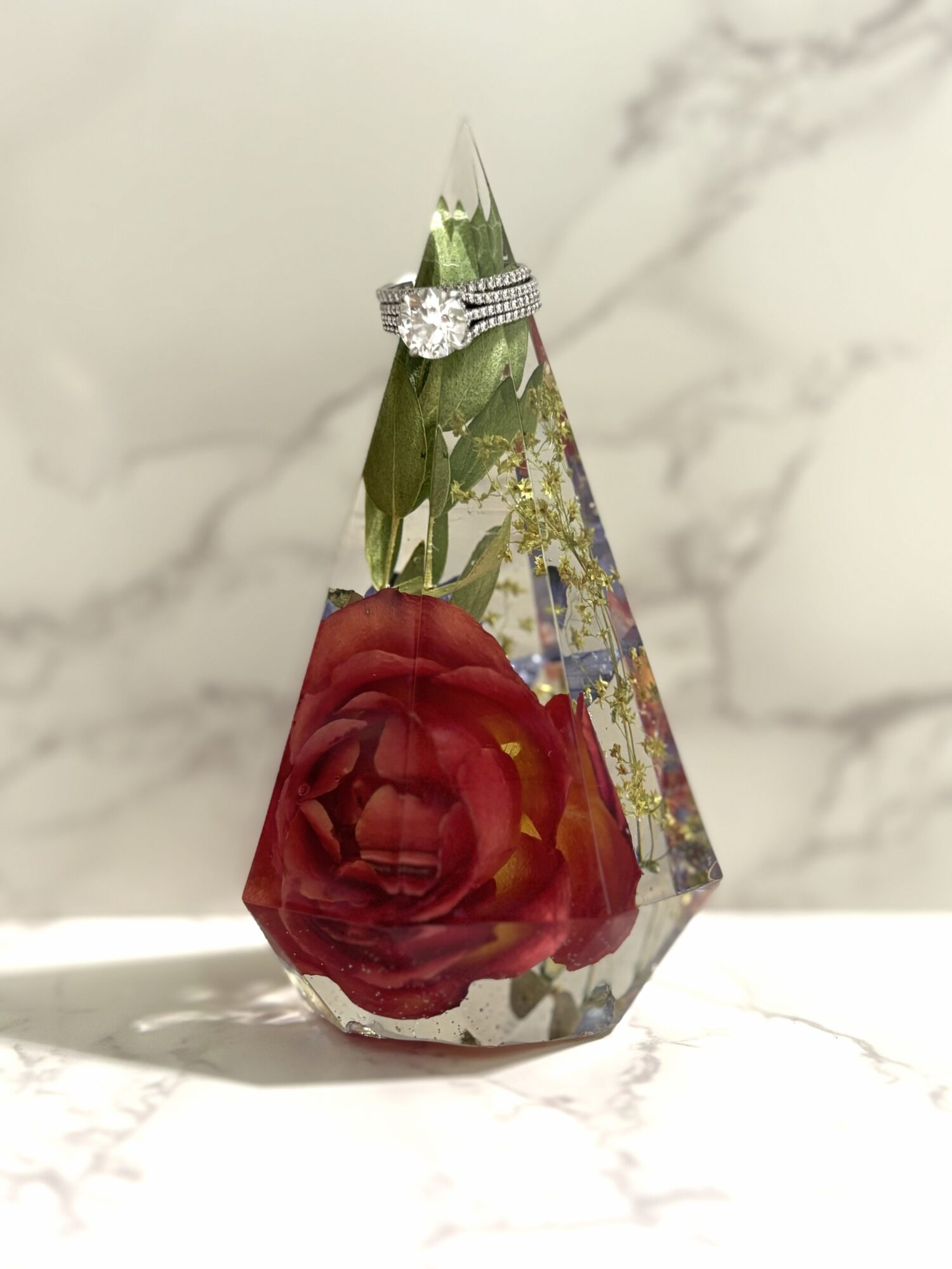 A teardrop-shaped glass ornament with a red flower inside, placed on a marble surface with a ring on top.