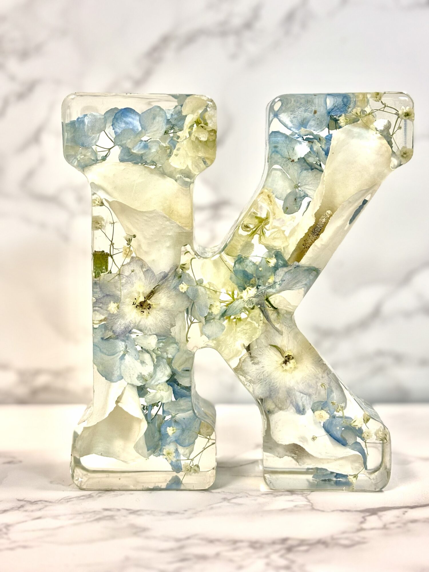Decorative letter K made of clear resin with embedded blue, white, and yellow flowers, placed on a marble surface.