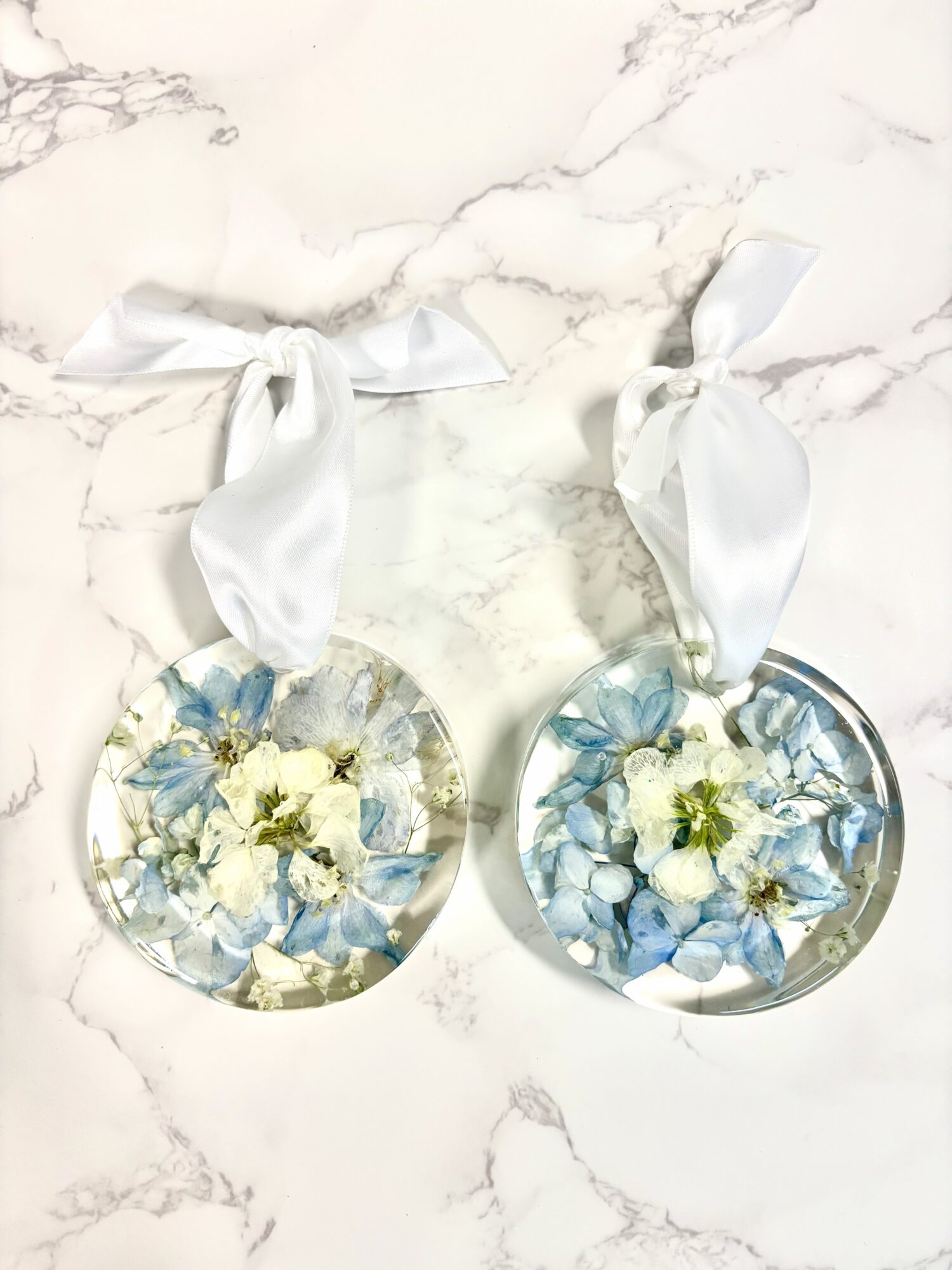 Two round floral ornaments with white ribbons on a marble surface, each containing white and blue flowers inside.