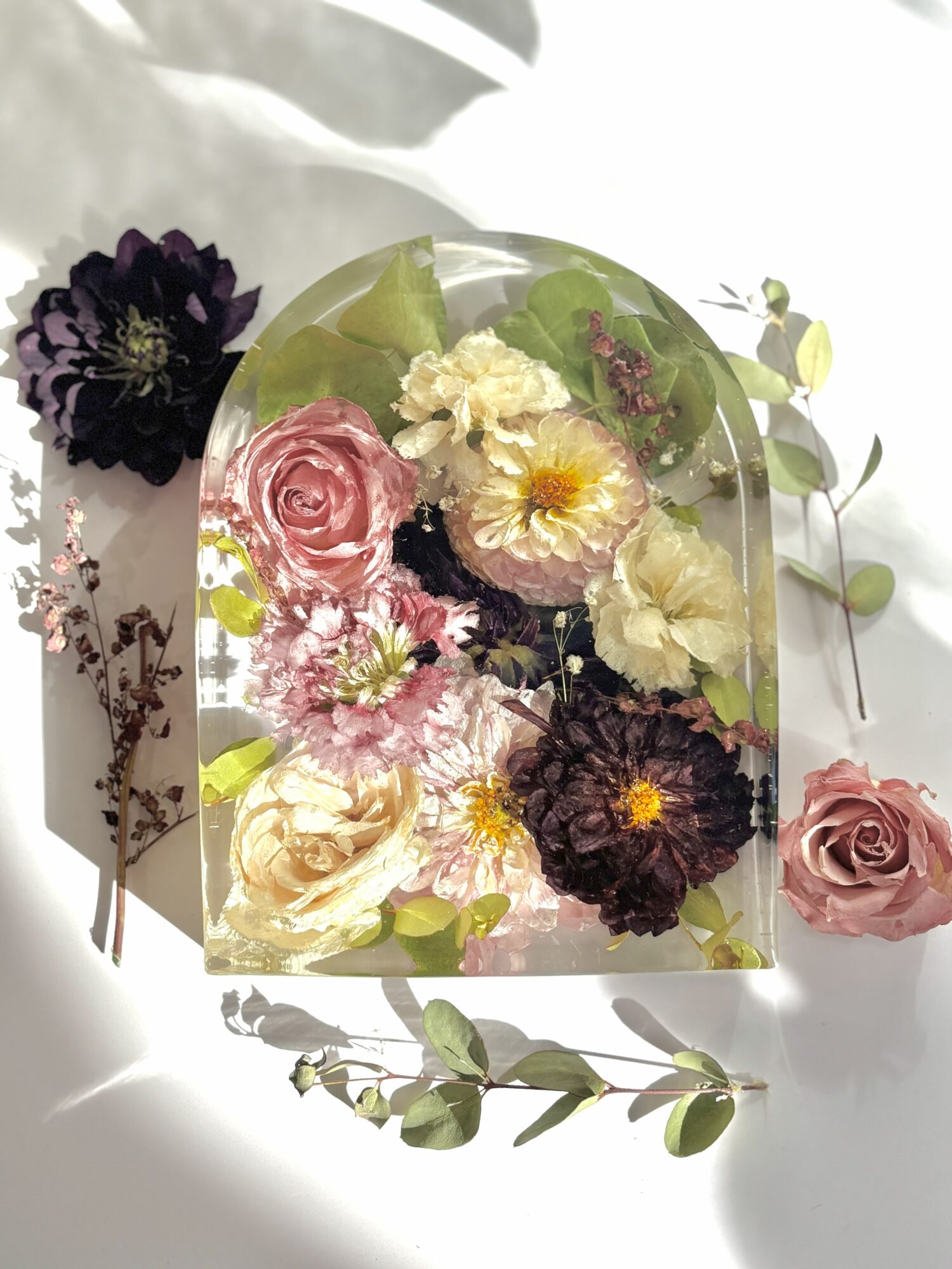 Arrangement of various flowers in a glass container with additional flowers and leaves around it, on a white surface.