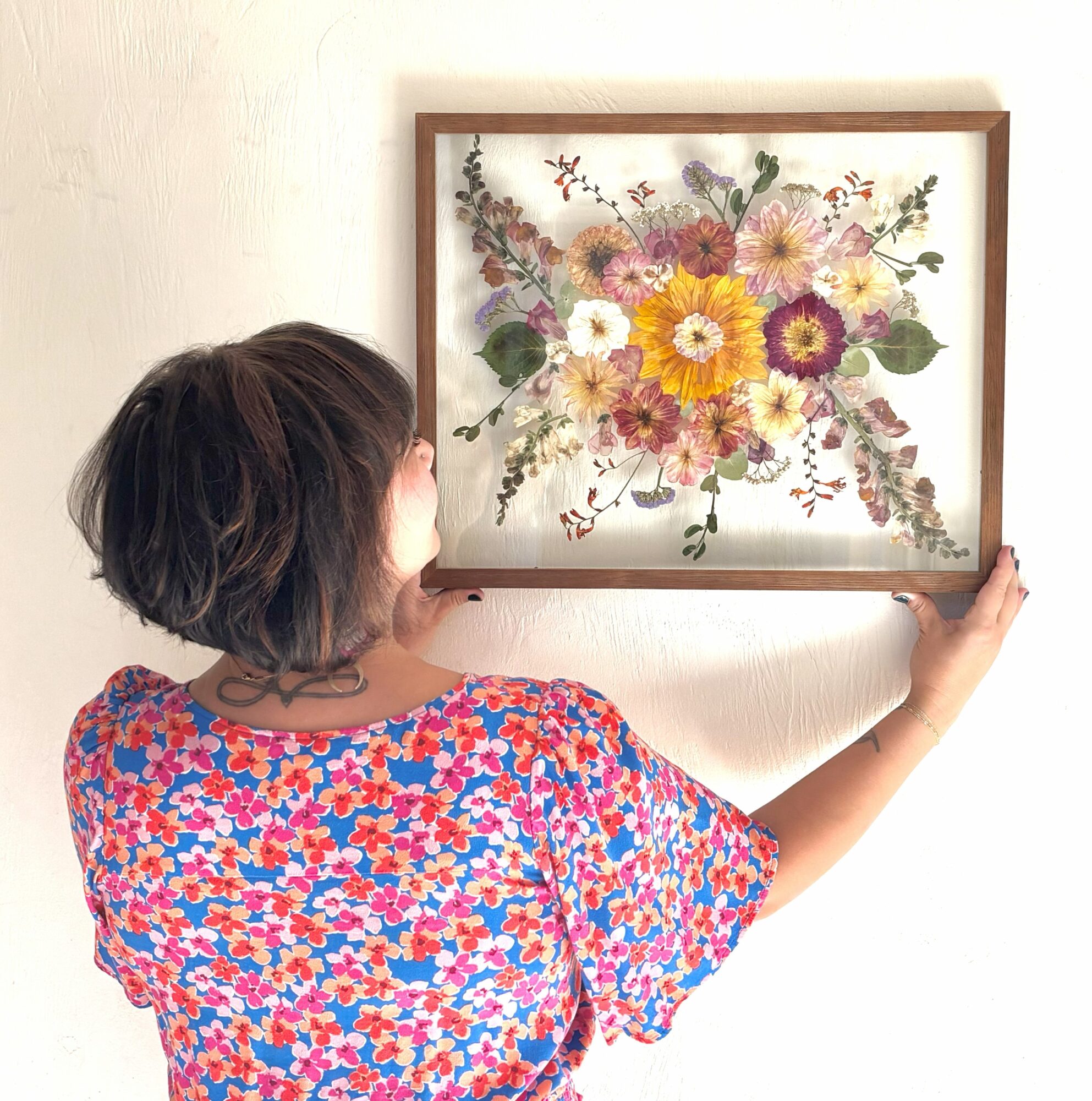 Child with short hair holding a framed floral painting on a wall, wearing a colorful patterned shirt.