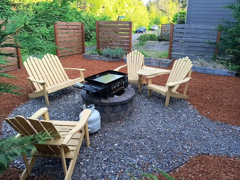 Outdoor seating area with four beige Adirondack chairs around a fire pit on gravel, surrounded by greenery and a blue building.