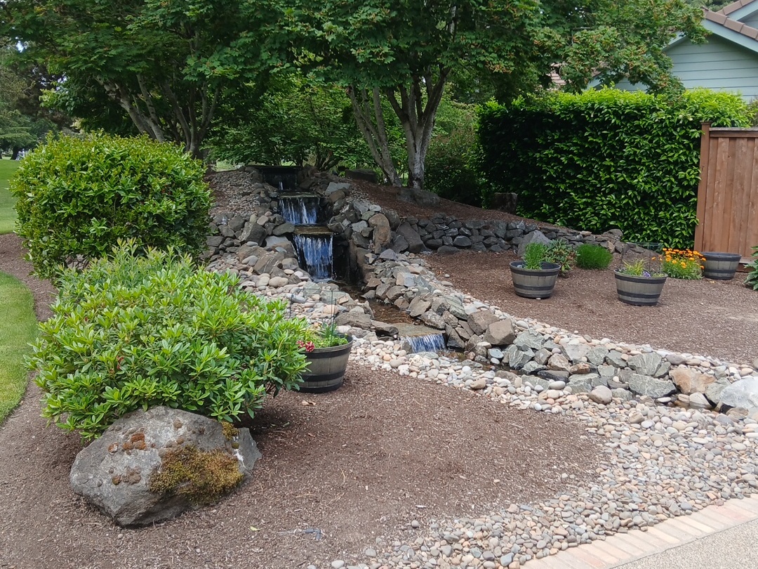 Garden with a small waterfall feature, rocks, potted plants, and green bushes, with trees and a house in the background.