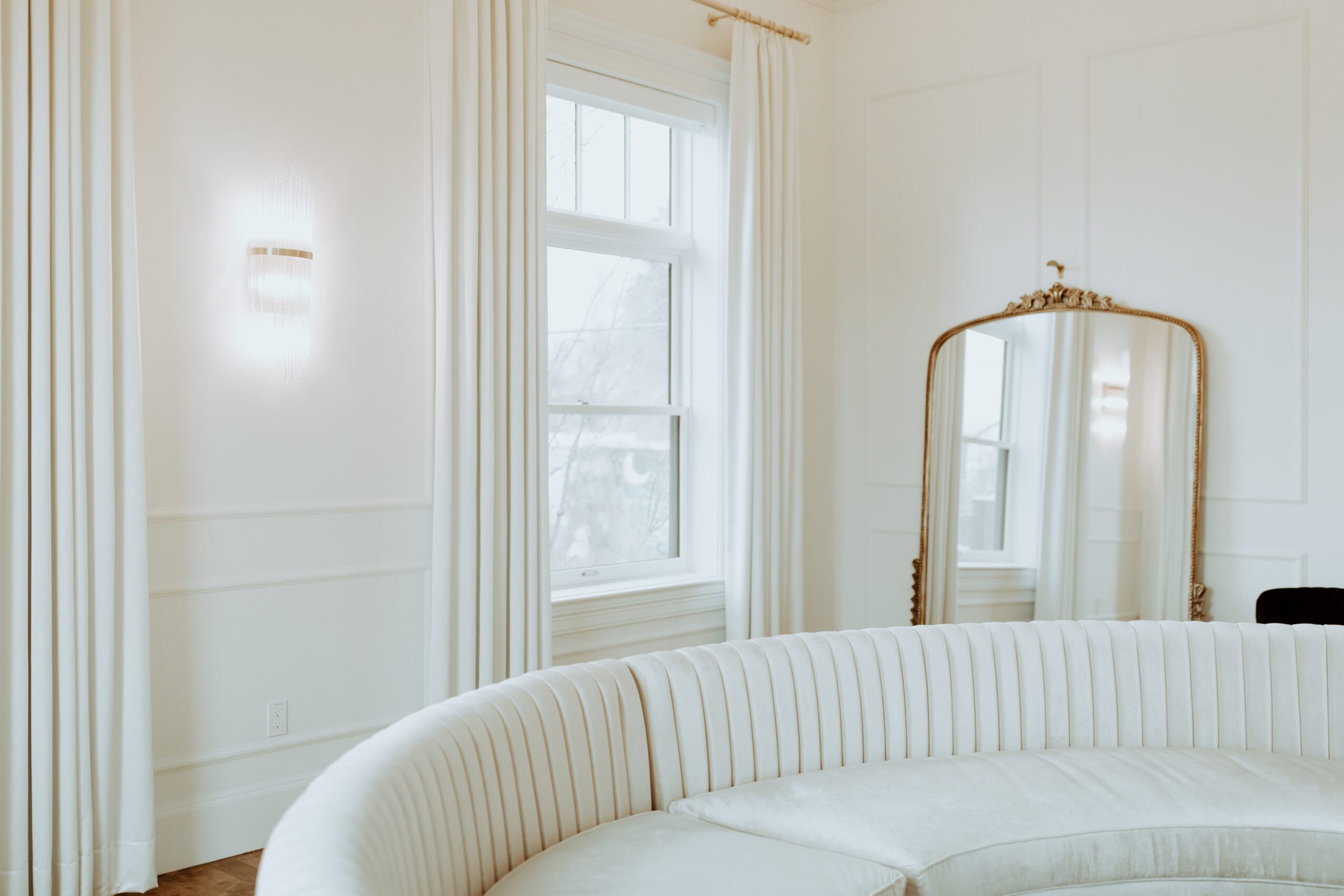 A bright room with a window, white curtains, a mirror, and a curved white sofa, with white walls and trim.