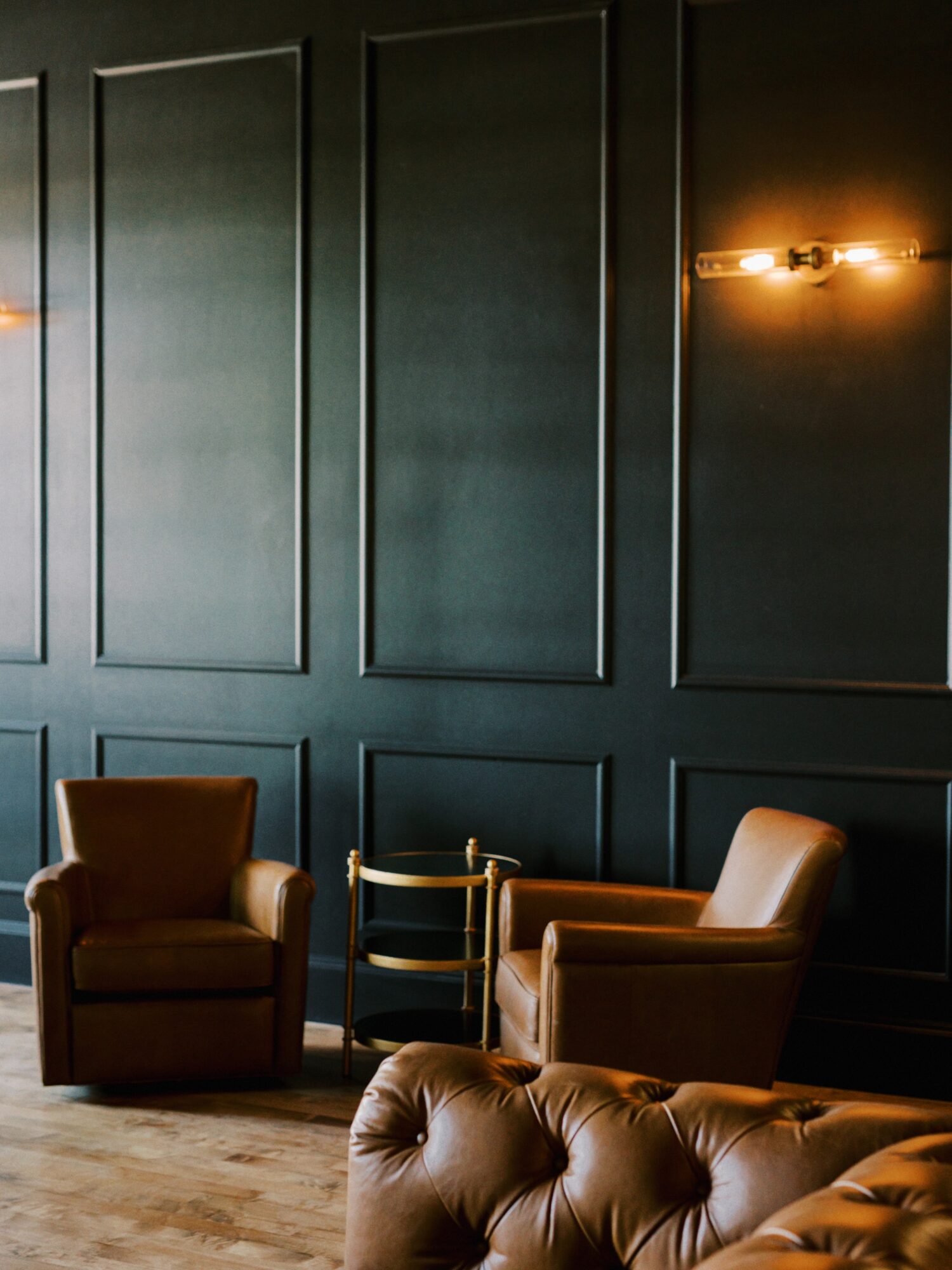 Two brown armchairs and a tufted sofa in a room with dark green paneled walls and warm wall sconces.