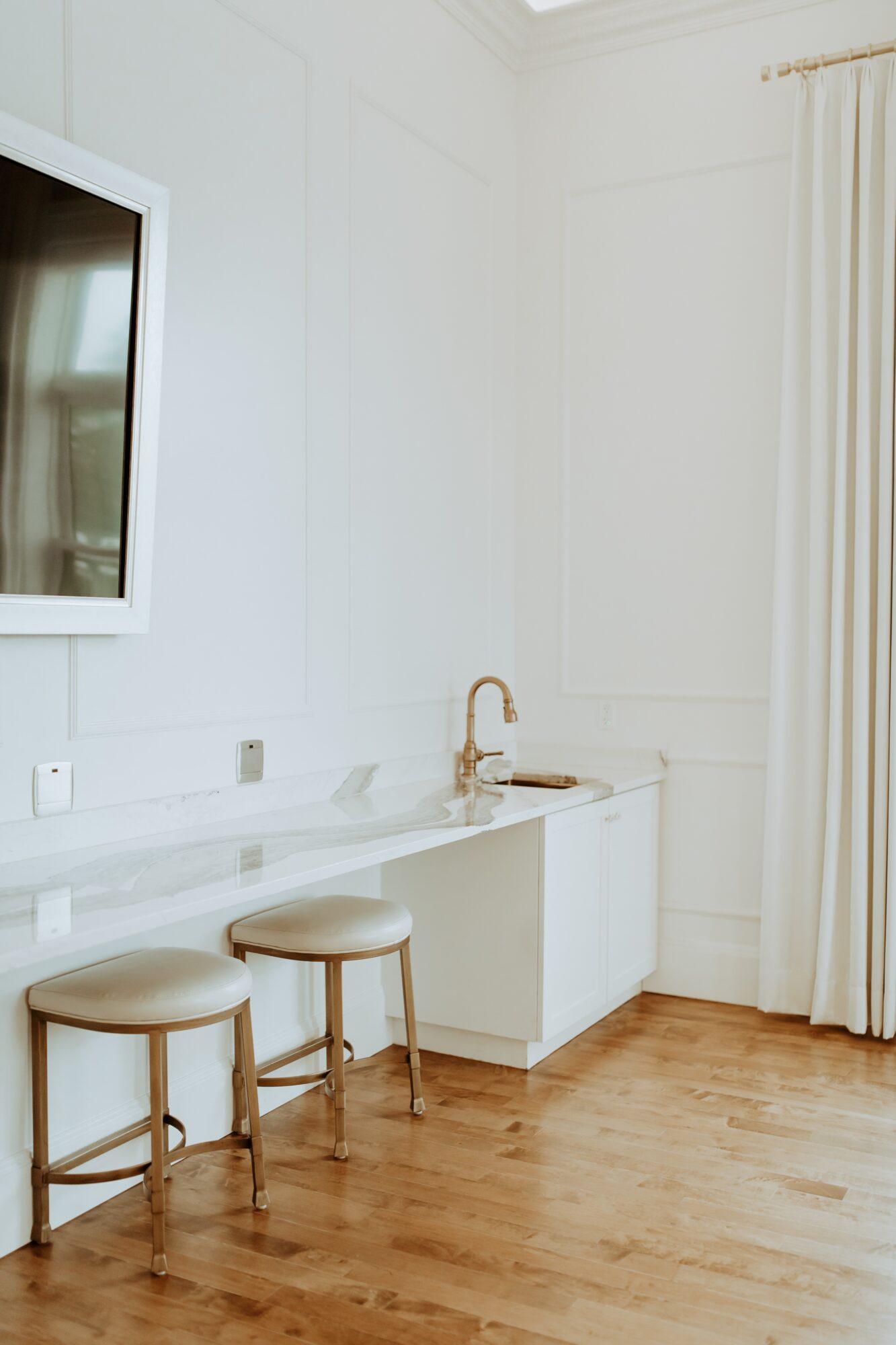 Corner of a room with a white countertop, a small sink, two stools, and a window with curtains.