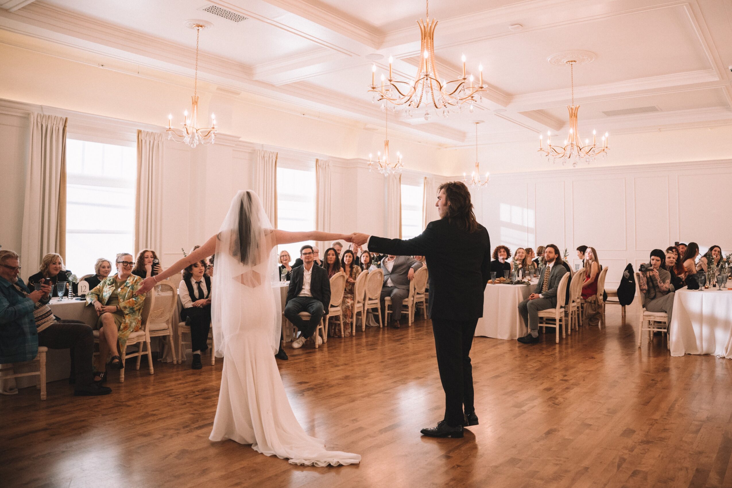 Bride and groom dancing in a decorated wedding hall with guests seated at tables, chandeliers overhead, and large windows with curtains.