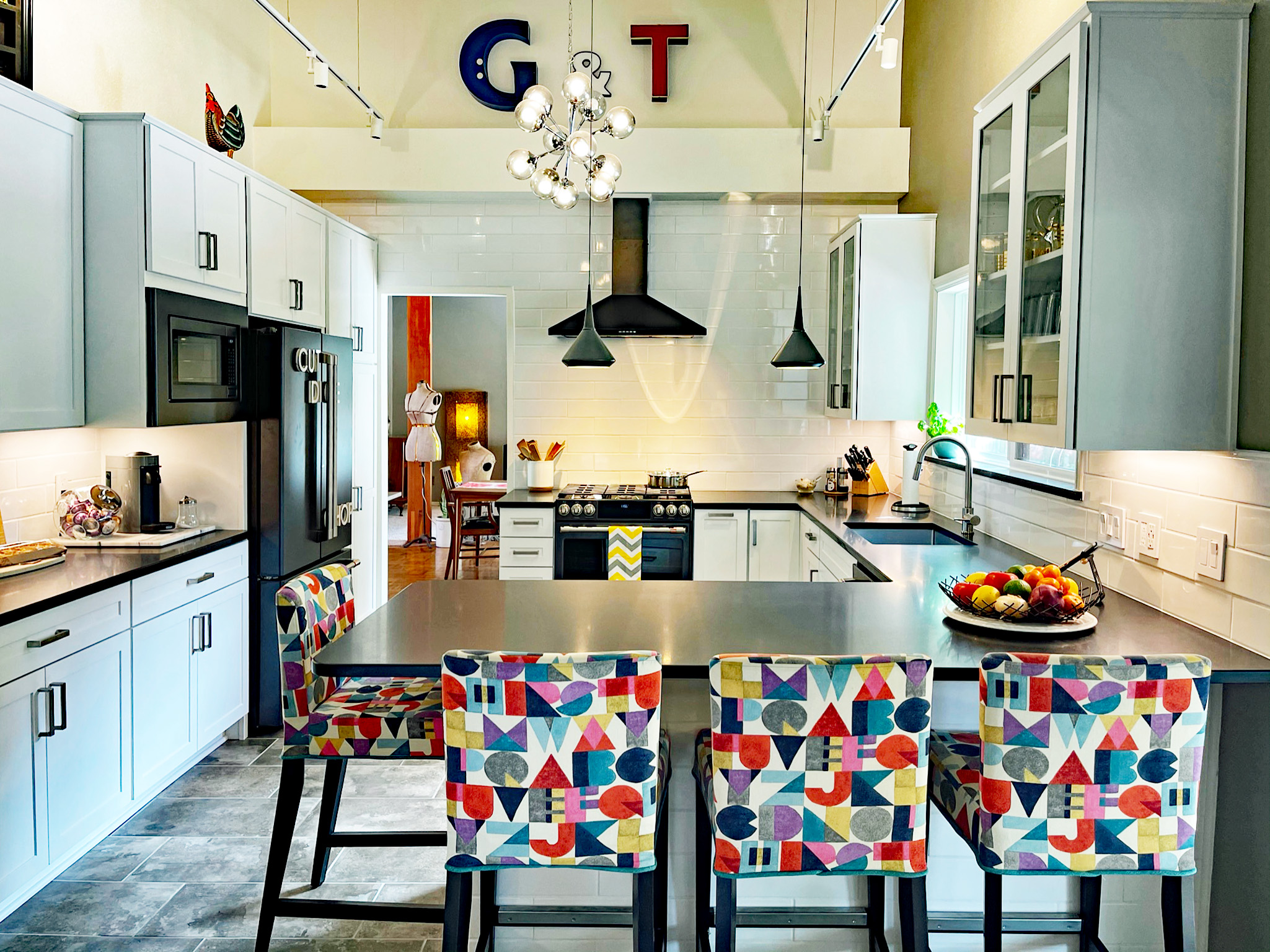Kitchen with light blue cabinets, a central island with colorful patterned chairs, and a modern range hood. Letters G and T hang from the ceiling.