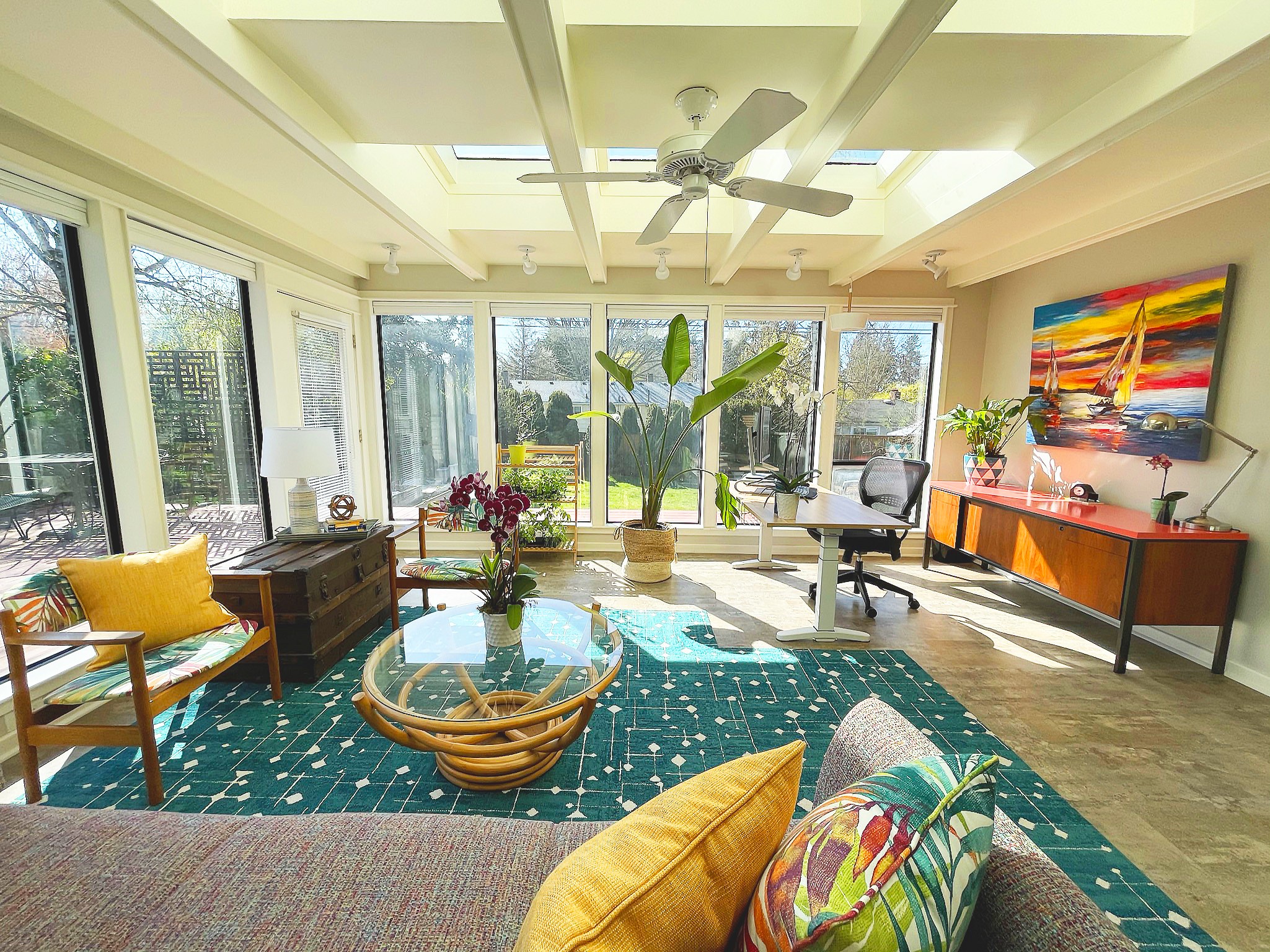 Bright living room with large windows, a ceiling fan, a green plant, colorful artwork, and a blue rug with yellow and patterned cushions.
