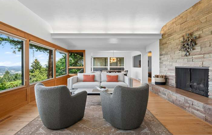 Living room with large windows, white sofa, two gray chairs, and a brick fireplace, with a view of trees outside.