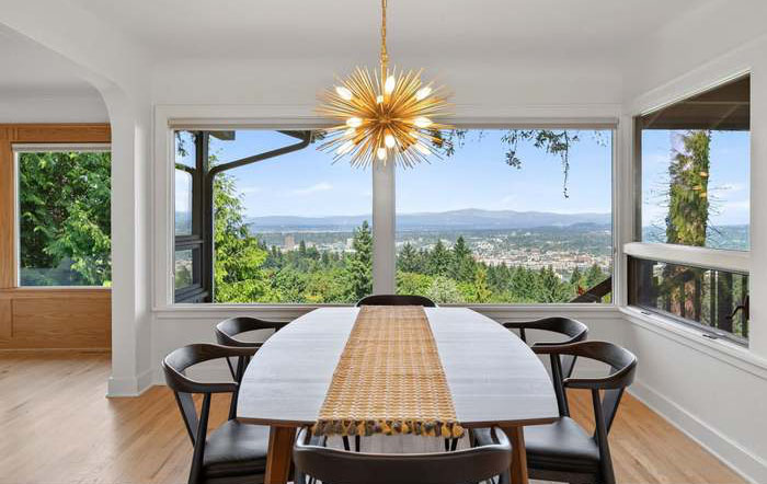 Dining room with large windows showing a landscape view, a white table with a woven runner, six chairs, and a starburst chandelier.