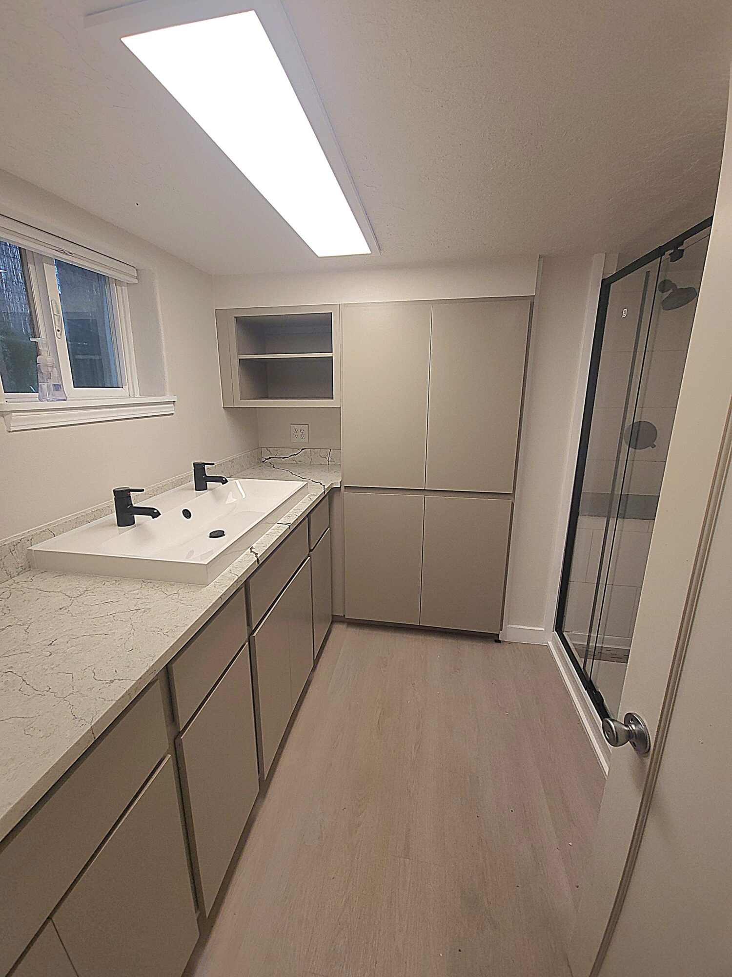 Bathroom with double sink, cabinets, window, and shower enclosure, illuminated by ceiling light, with light-colored flooring and walls.