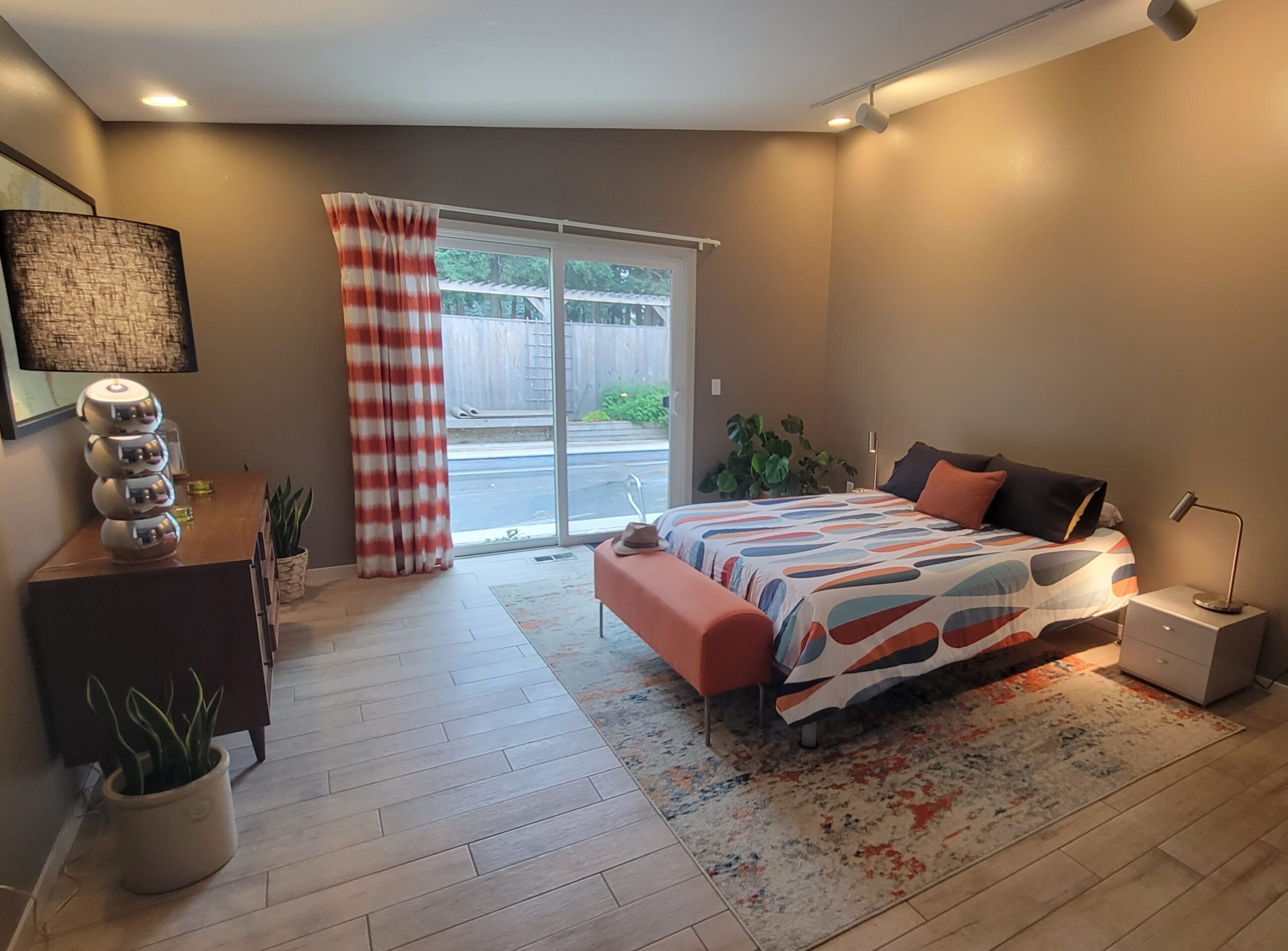 Bedroom with bed, sliding glass door, curtains, dresser, plants, and rug, illuminated by ceiling and side lamps.