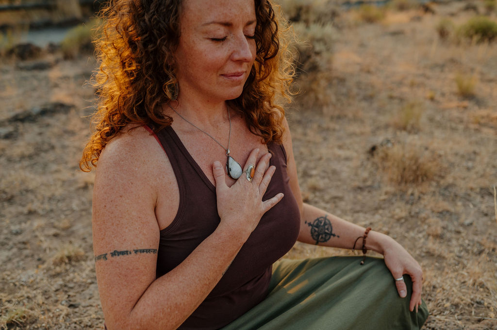 Woman with curly hair sitting outdoors, eyes closed, hand on chest, wearing a dark top and green skirt.