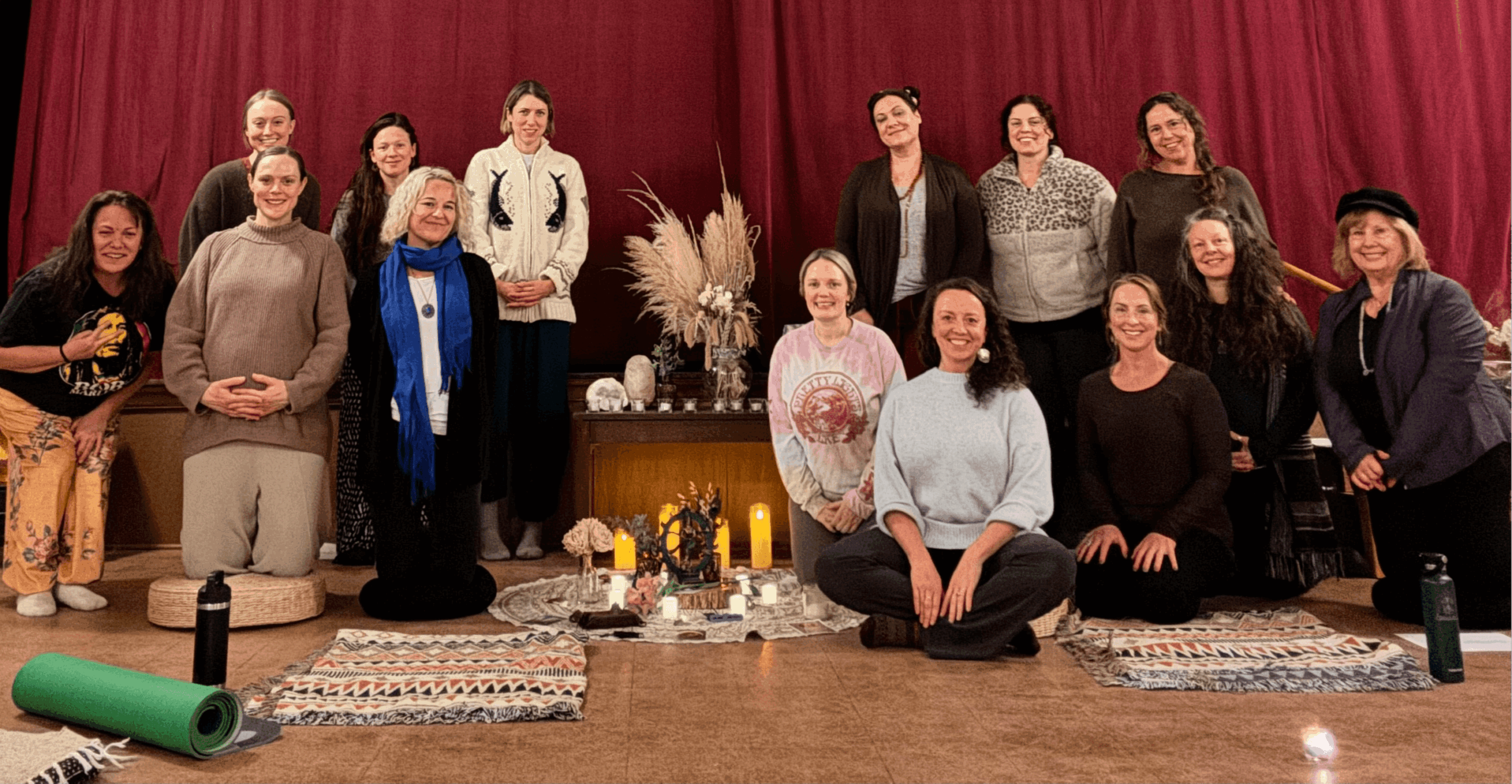 Group of 14 people in a room with red curtains, some sitting on the floor, others standing, with decorative items and candles.