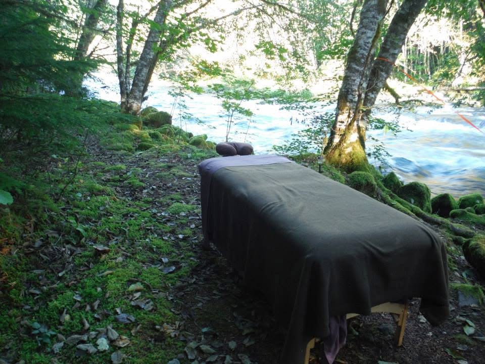 A massage table with a black cover is set up outdoors near a river, surrounded by trees and greenery.