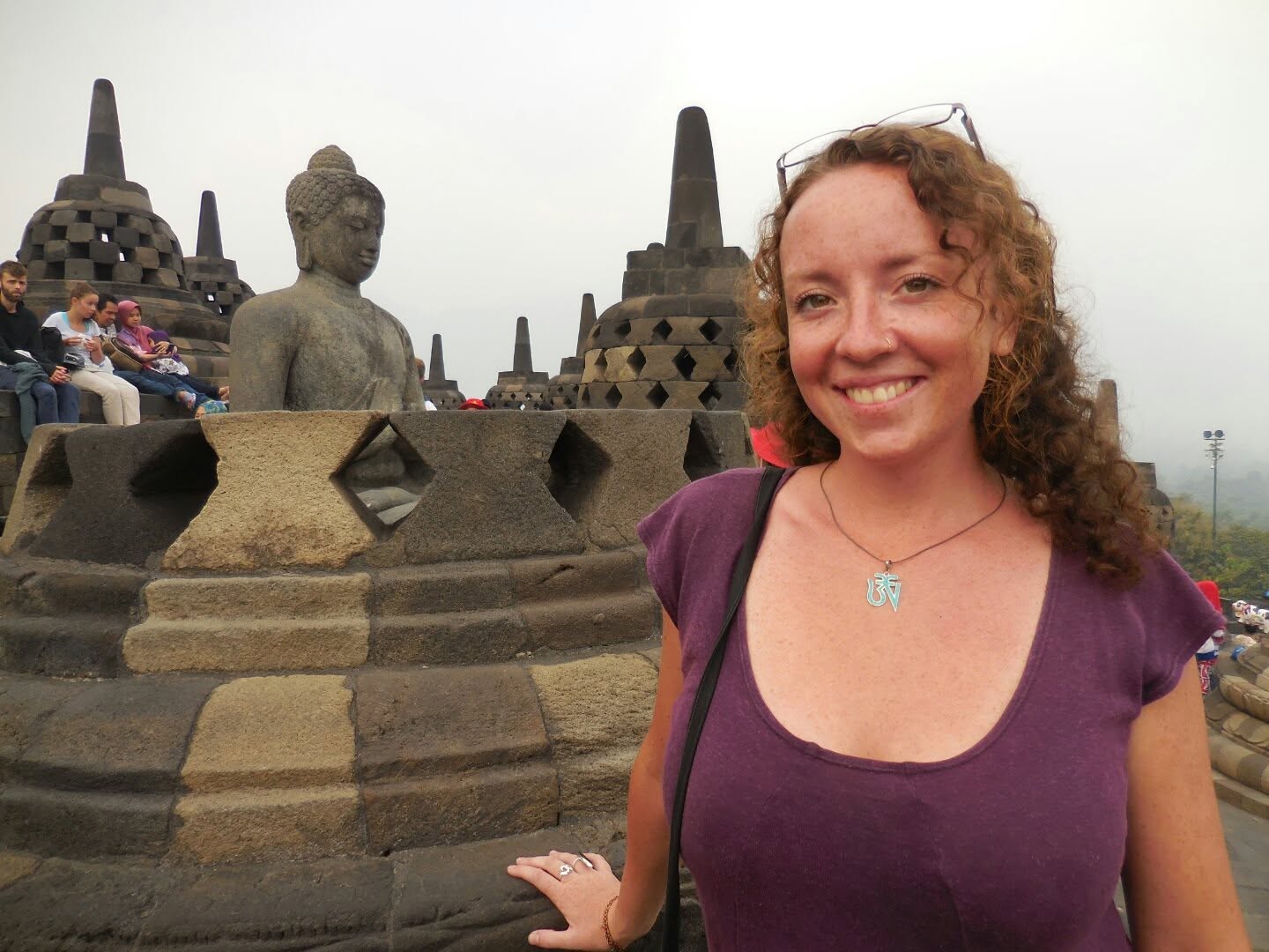 Smiling woman with curly hair standing near ancient stone structures and statues, cloudy sky background.