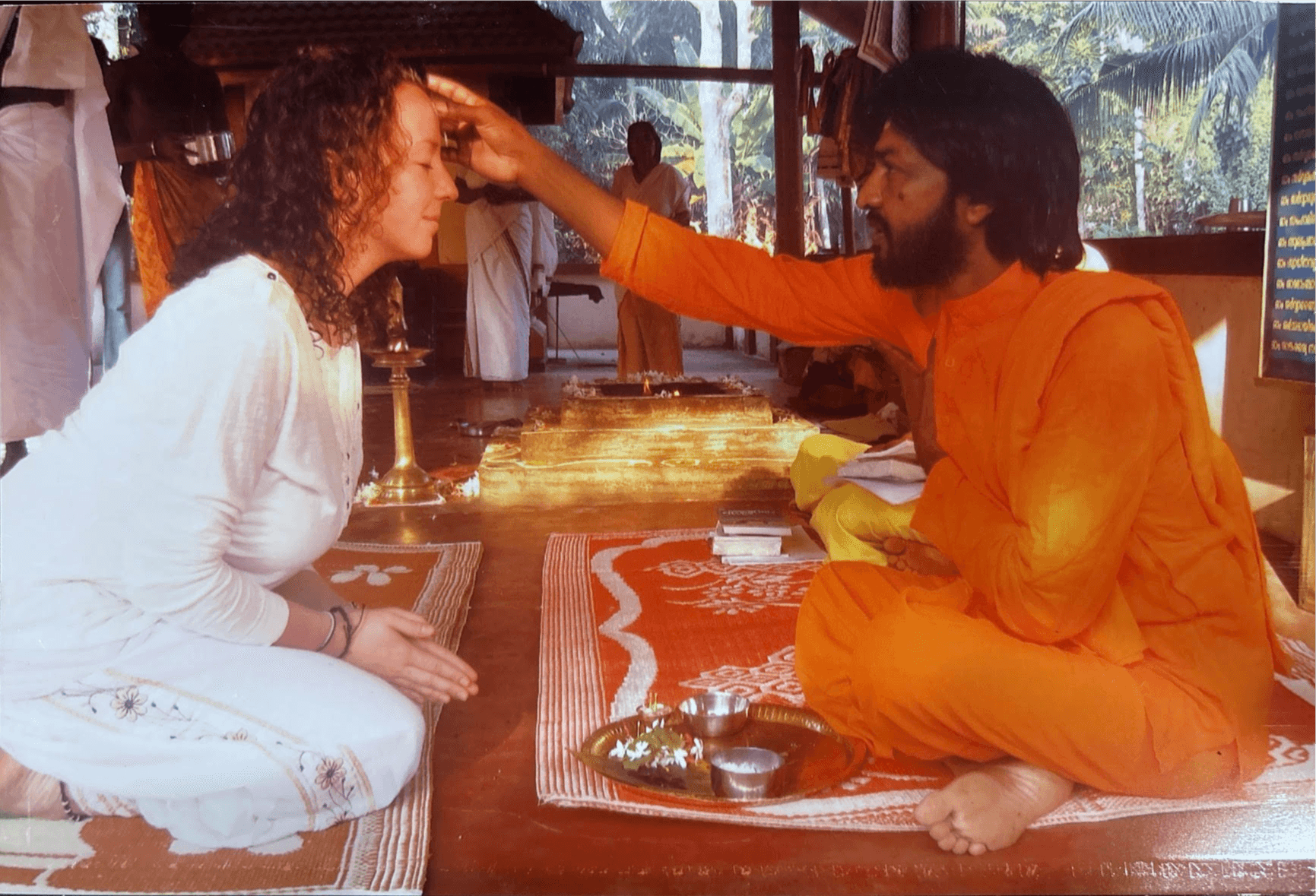 A person in orange robes performs a blessing or ritual on a woman in white, inside a temple or spiritual setting.