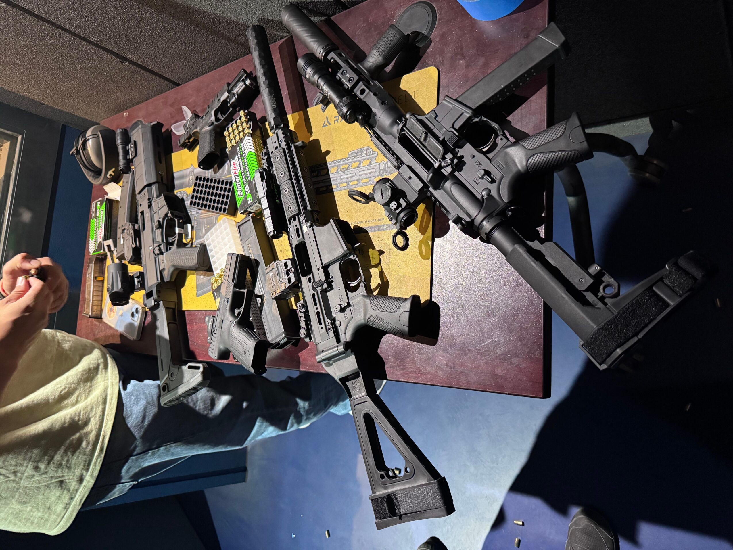 Three black rifles with scopes and attachments on a yellow surface, with a person's hand and arm visible.