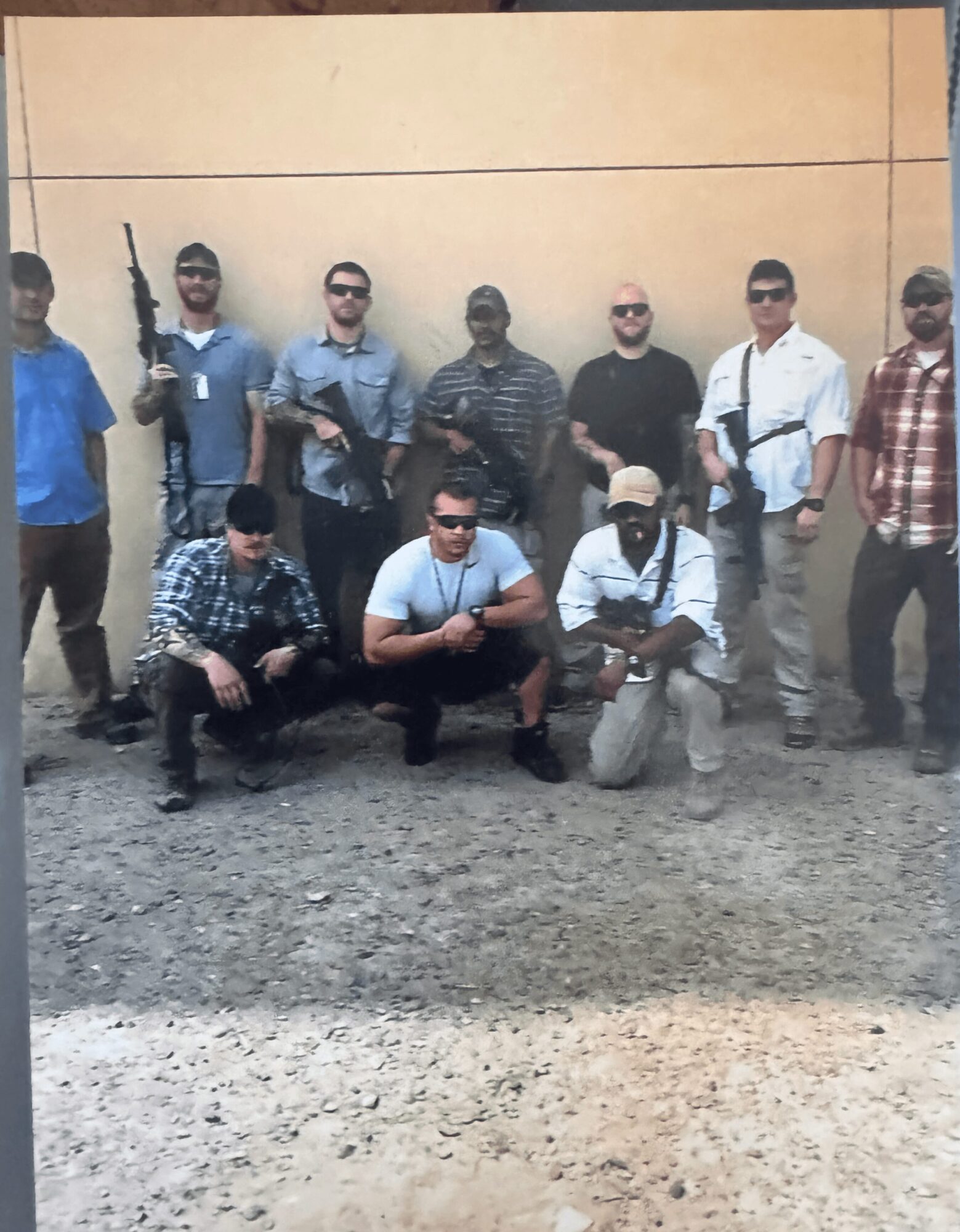 Group of ten men standing and kneeling outdoors against a beige wall, some holding weapons, casual clothing, gravel ground.