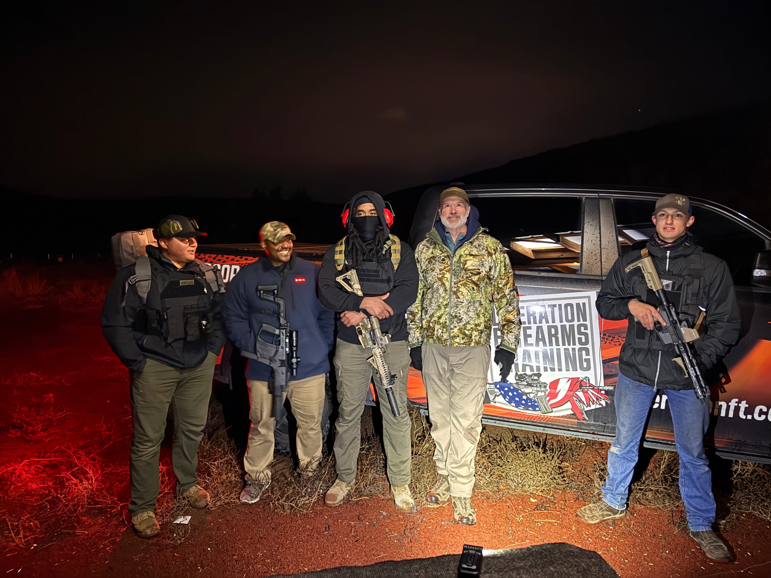 Group of six people standing outdoors at night near a vehicle with a sign, some wearing tactical gear and masks.