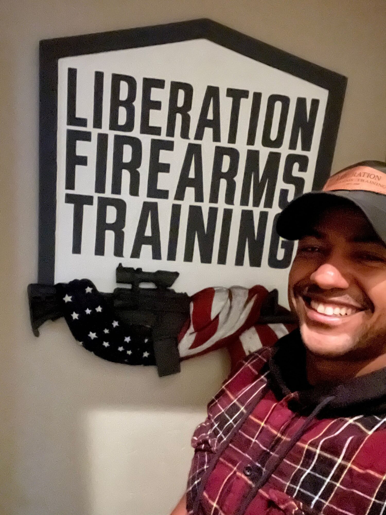 Person smiling, wearing a cap and plaid shirt, in front of a sign that reads 'LIBERATION, FIREARM TRAINING'.