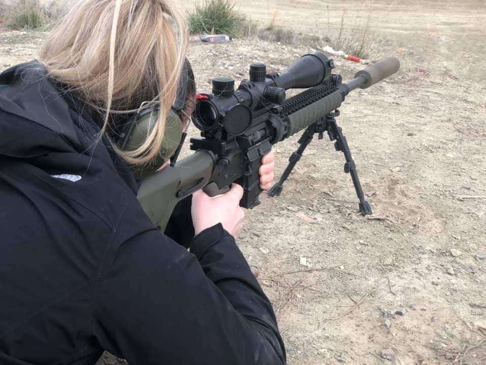 Person aiming a scoped rifle with bipod on dry, open ground, wearing a black jacket and glasses.