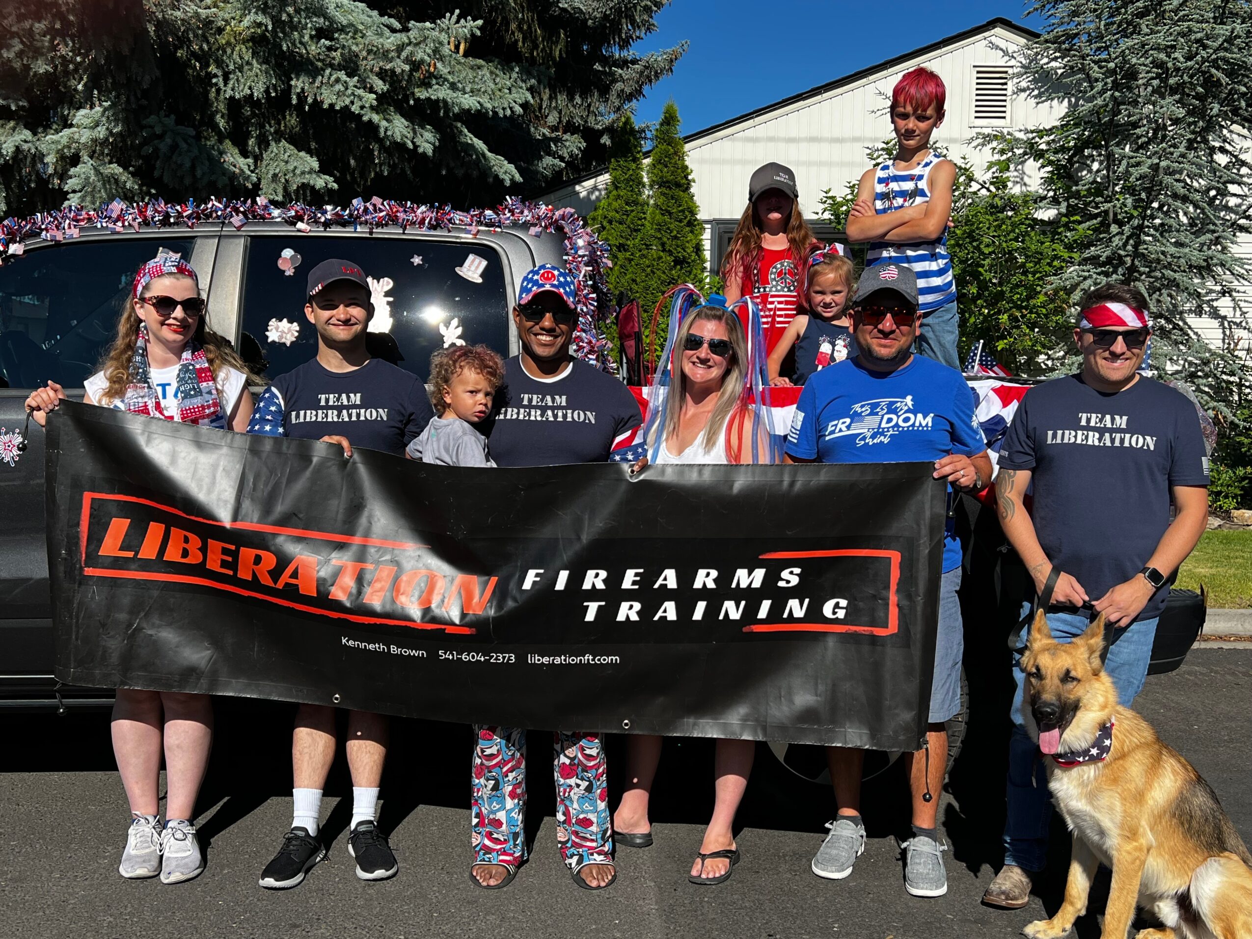 Group of people holding a banner outdoors, with a dog sitting nearby, in front of a house and trees.