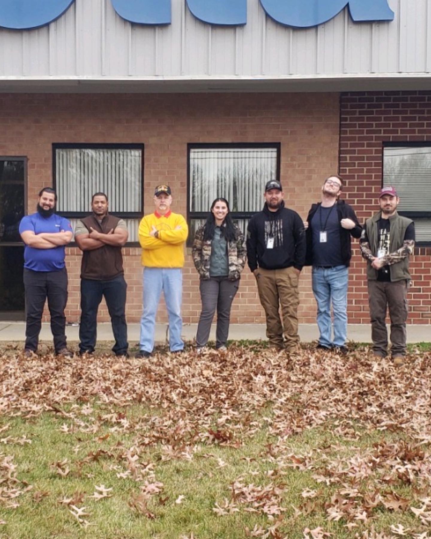 Seven people stand outside a building with windows and a brick wall, on a ground covered with fallen leaves.