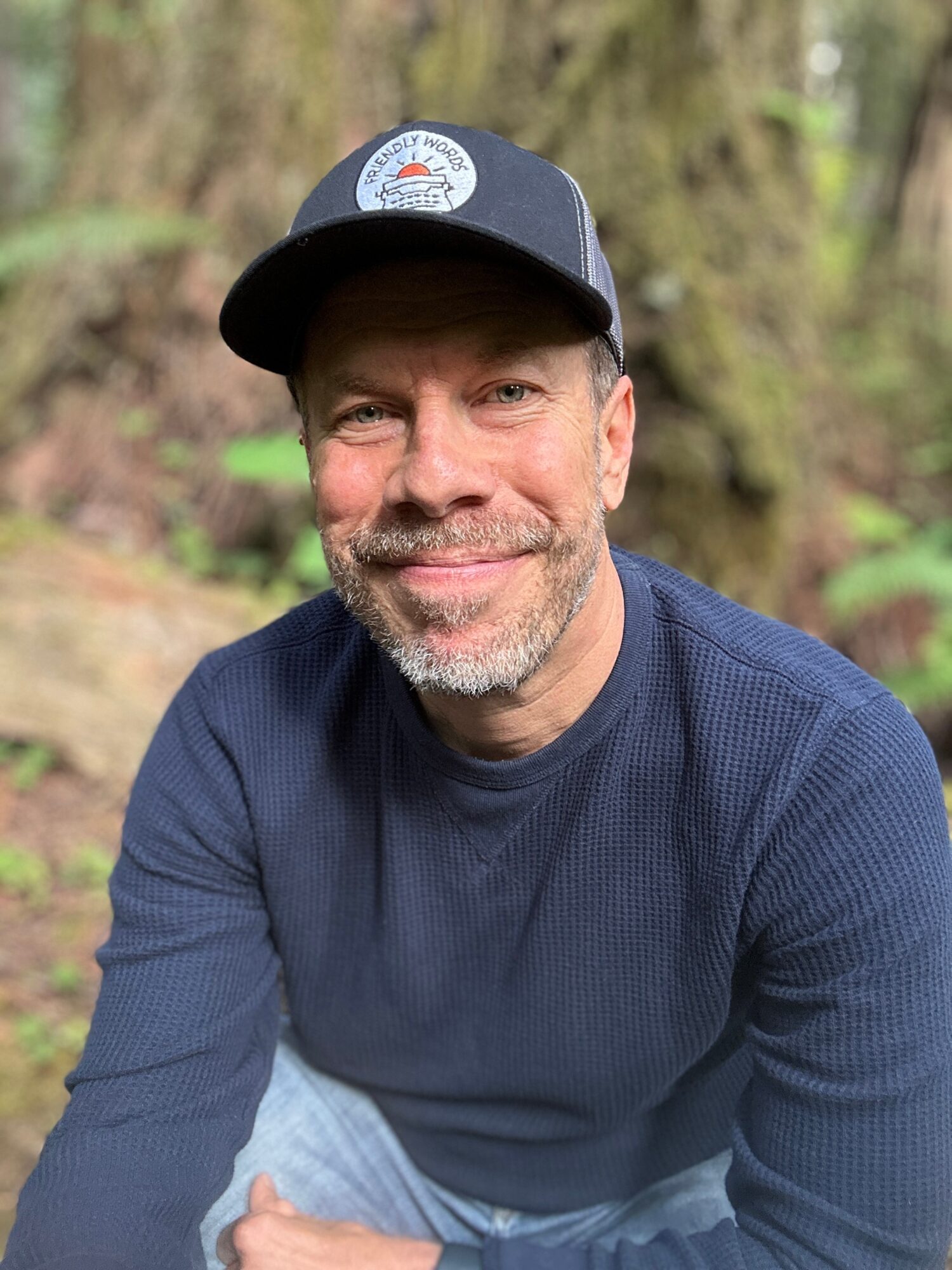Smiling man outdoors wearing a baseball cap and navy sweater, with a blurred forest background.