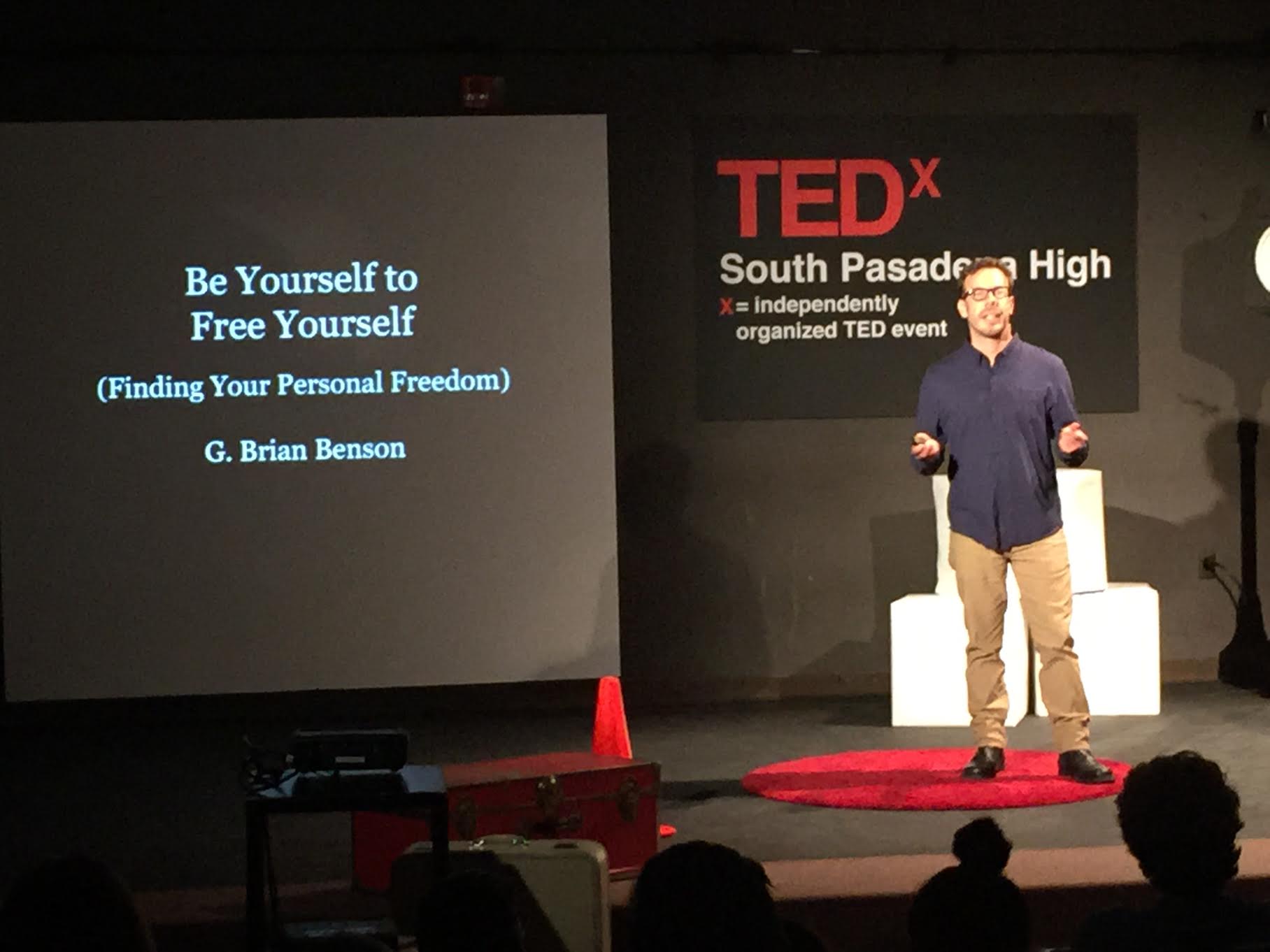 Speaker on stage at TEDx event, presentation slide titled 'Be Yourself to Free Yourself' by G. Brian Benson, audience in foreground.