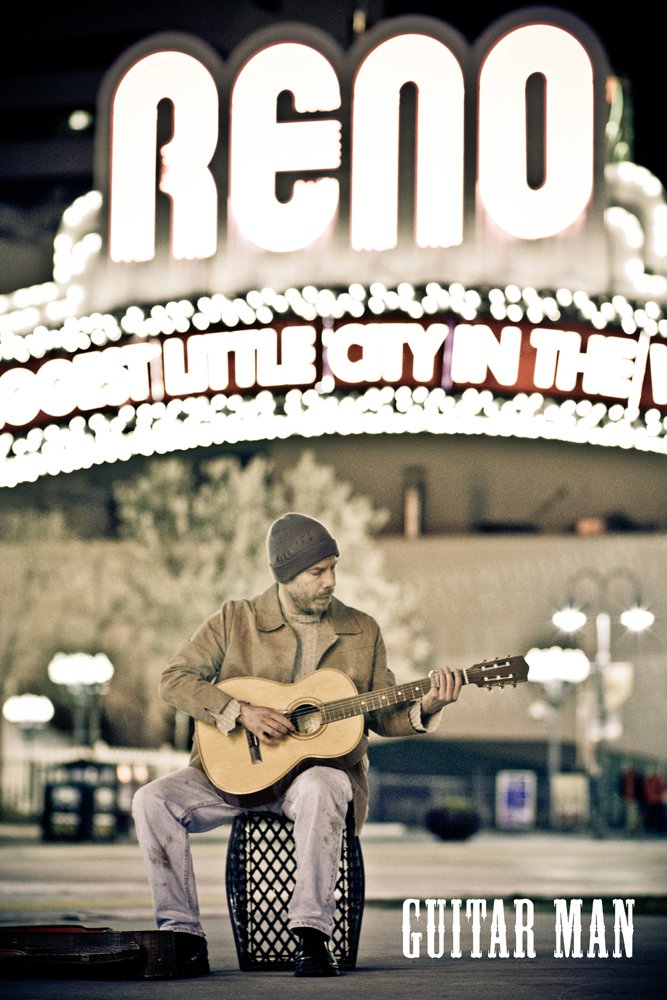 Person sitting outdoors playing guitar at night with illuminated sign reading 'RENO' in background.