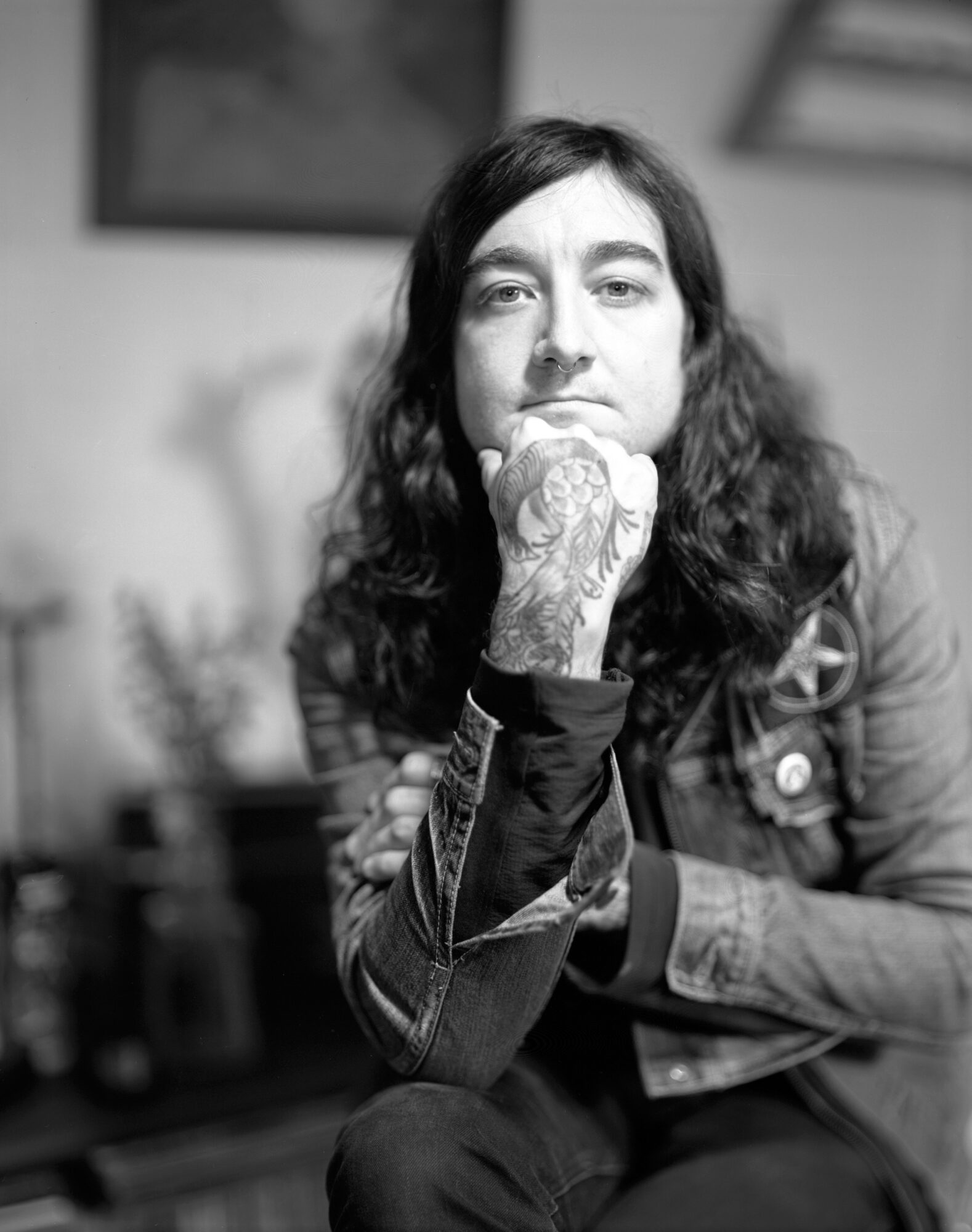 Black and white photo of a person with long hair, resting chin on hand, with tattoos visible on hand, in an indoor setting.